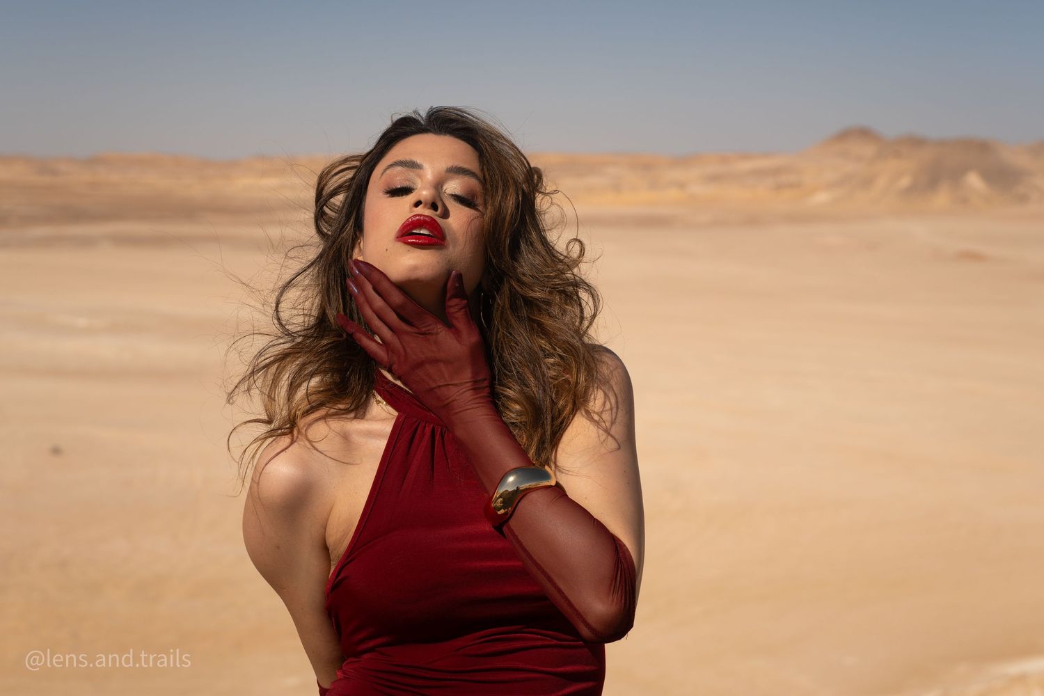 Woman in red dress with gloves tilts her head back gracefully in a sandy desert.