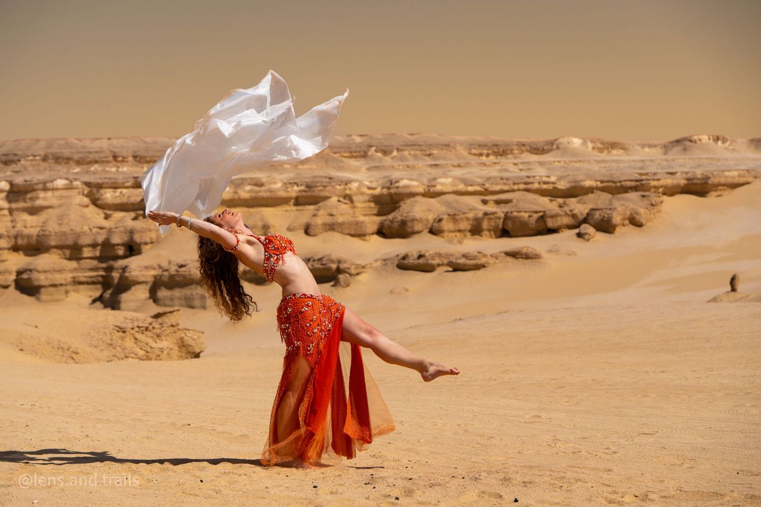 Belly dancer in red costume waves white fabric veil in arid desert landscape with rocky formations.