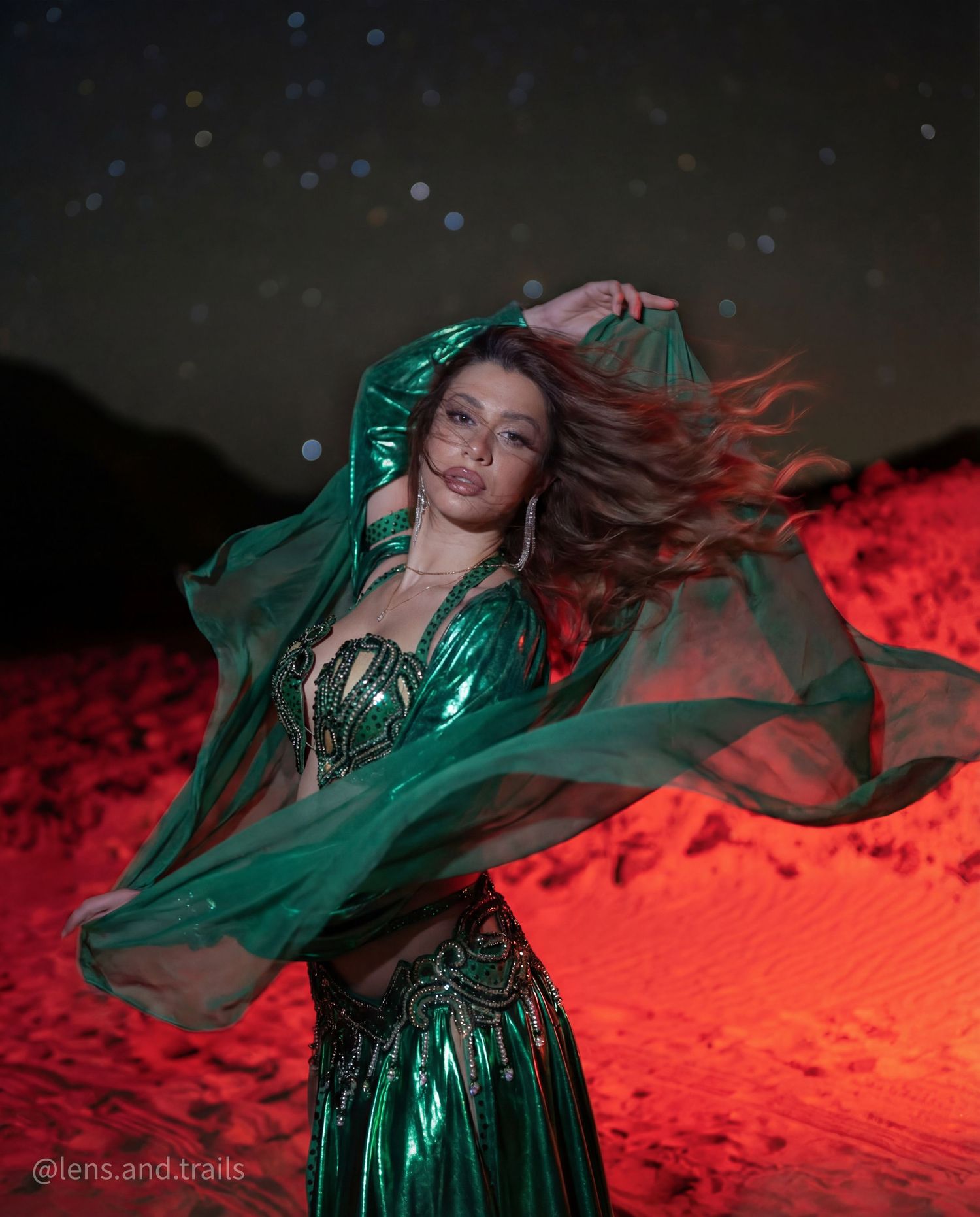 Belly dancer in emerald green costume performs dramatically at night in red-lit desert landscape.