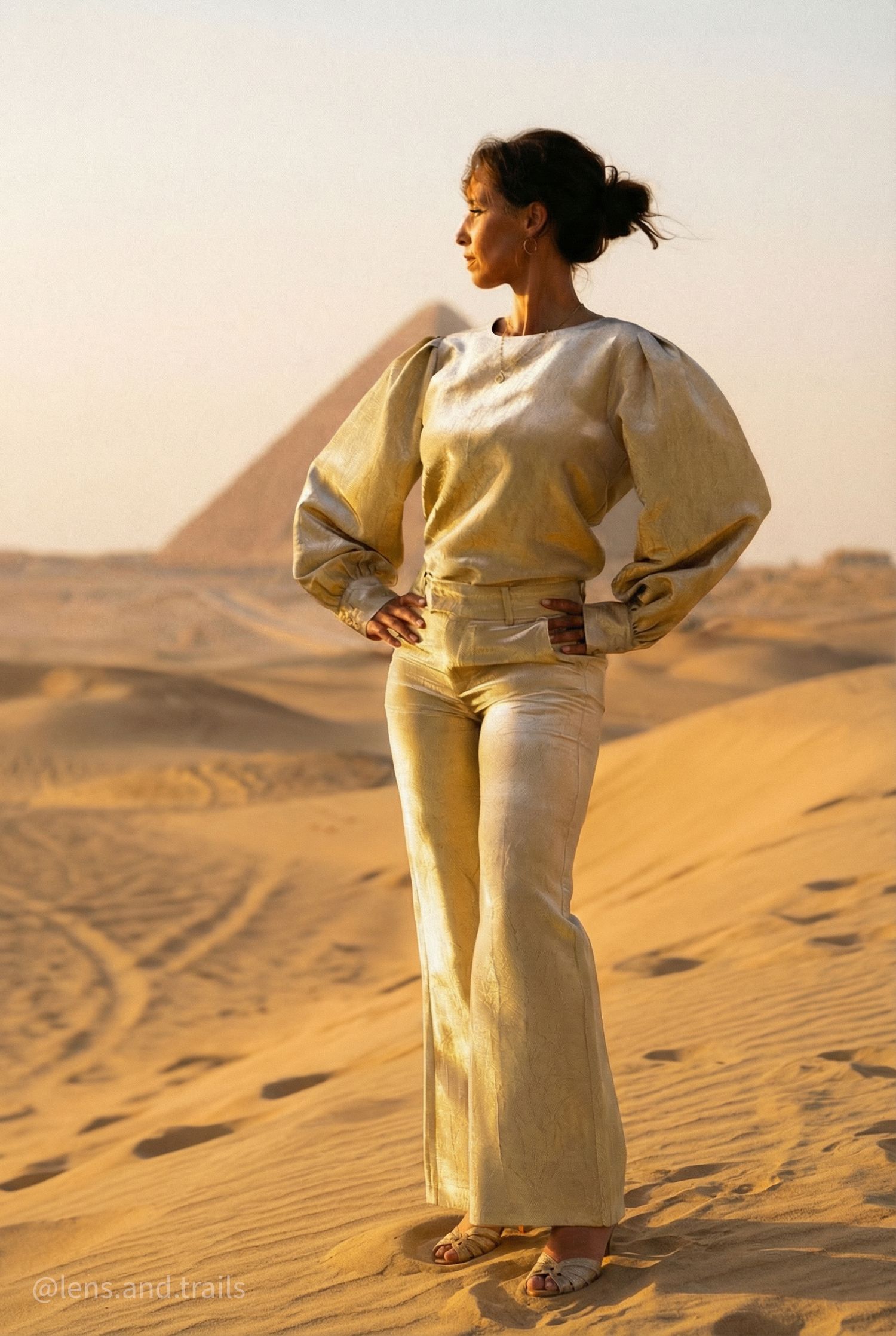 Woman in gold outfit stands confidently on desert sand with Egyptian pyramid in background.