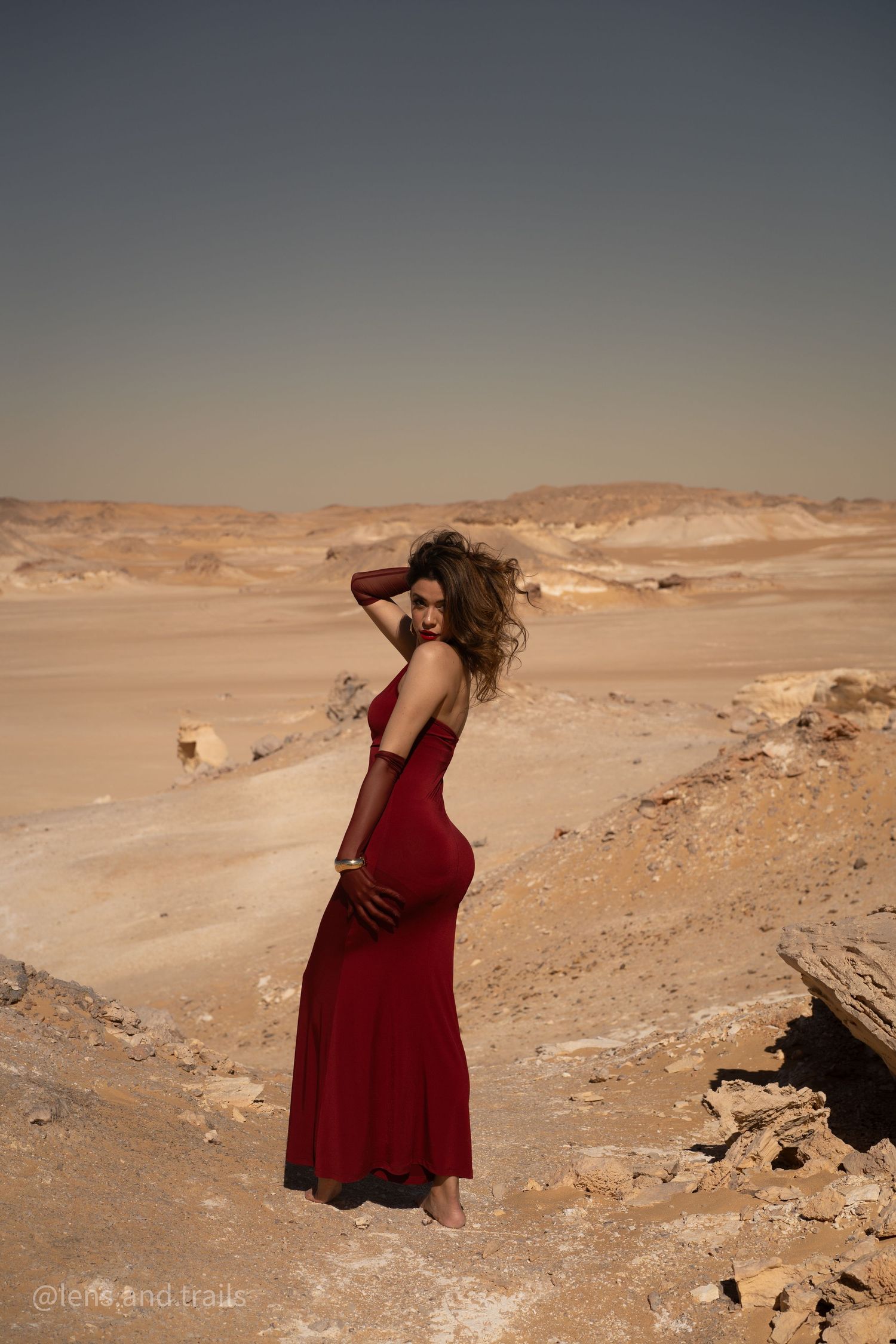 Woman in elegant red gown standing in vast desert landscape, looking back over her shoulder.
