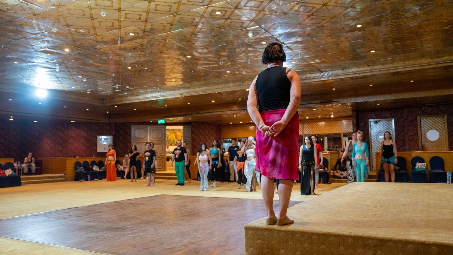 Dancer in red skirt stands barefoot on a wooden floor in a large ornate ballroom.
