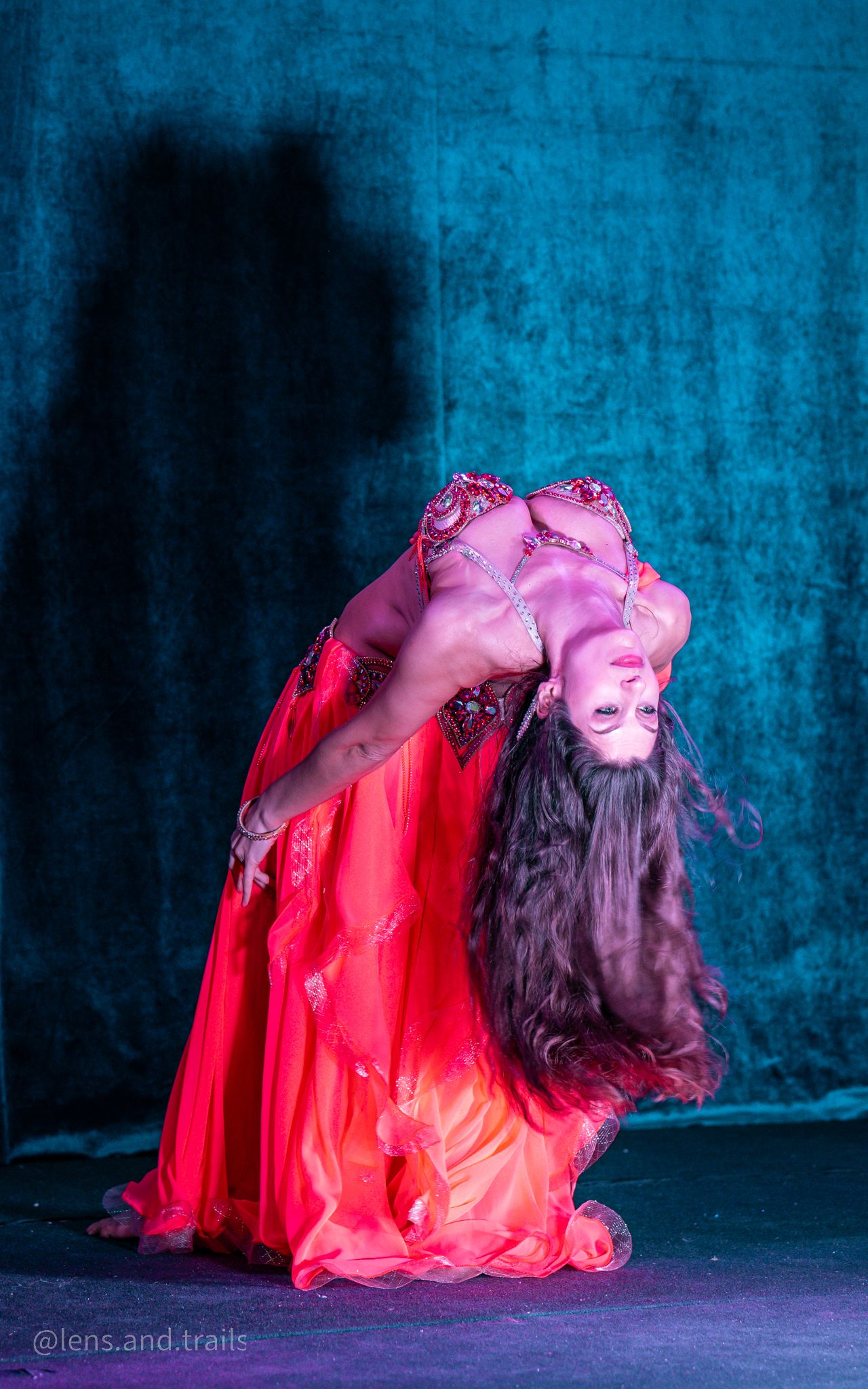 Belly dancer in red costume executing a deep backbend with hair flowing to the floor on stage.