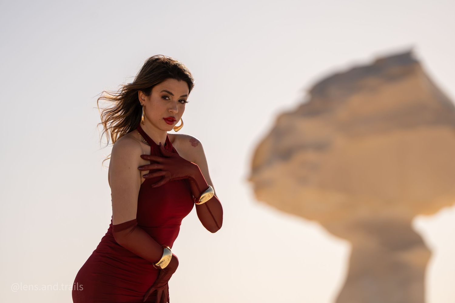 Woman in red dress poses with blurred mushroom-shaped desert rock formation behind her.