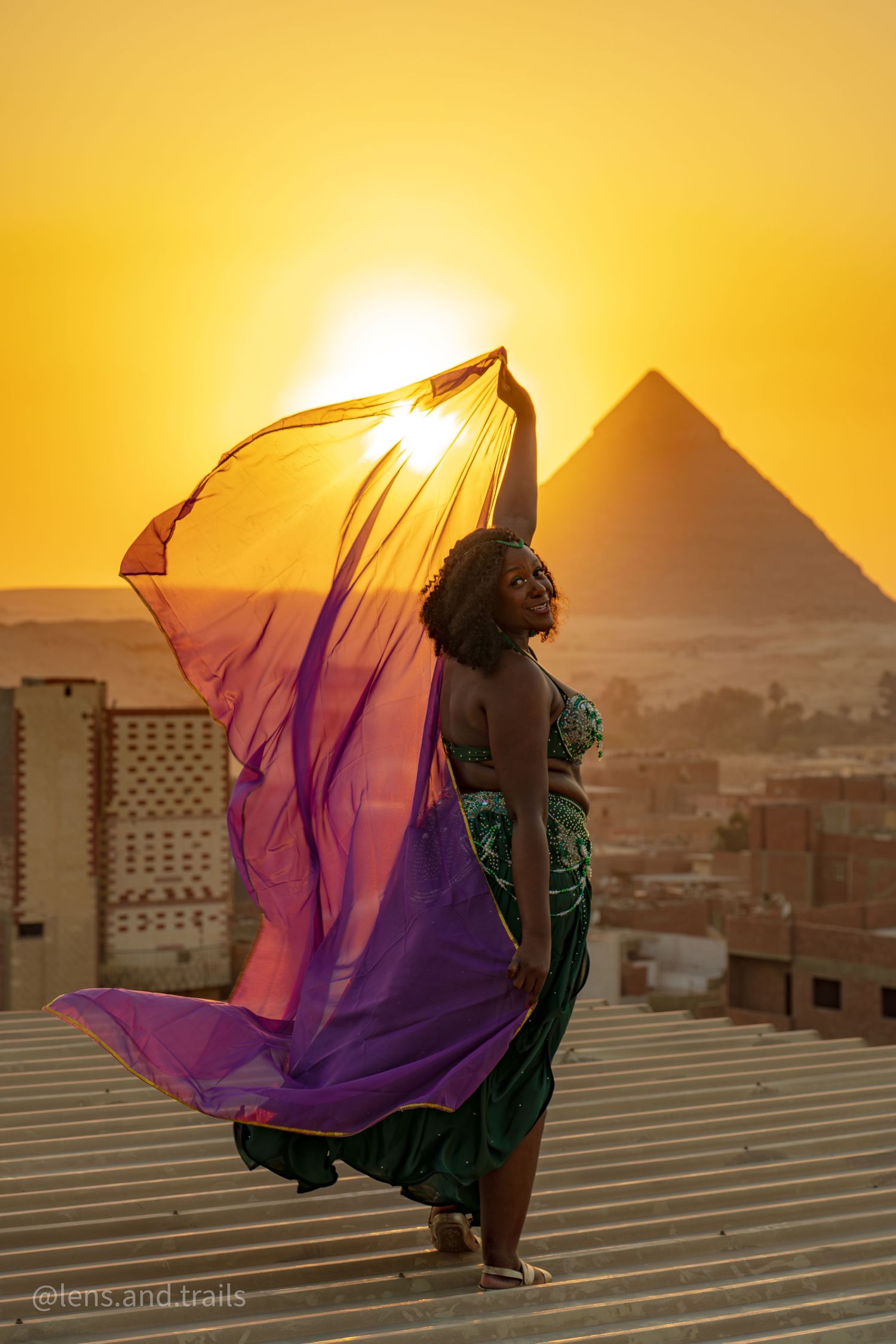 Woman in green dress waves flowing purple fabric before the Great Pyramid during a golden sunset.