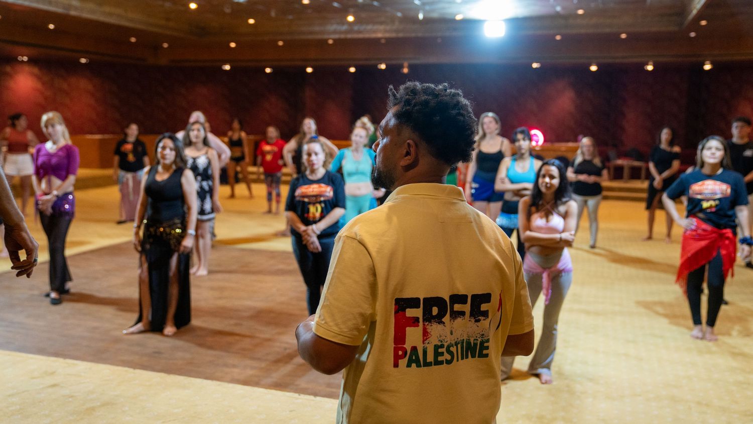 Dance instructor wearing Free Palestine shirt leads belly dance workshop in large studio with students.