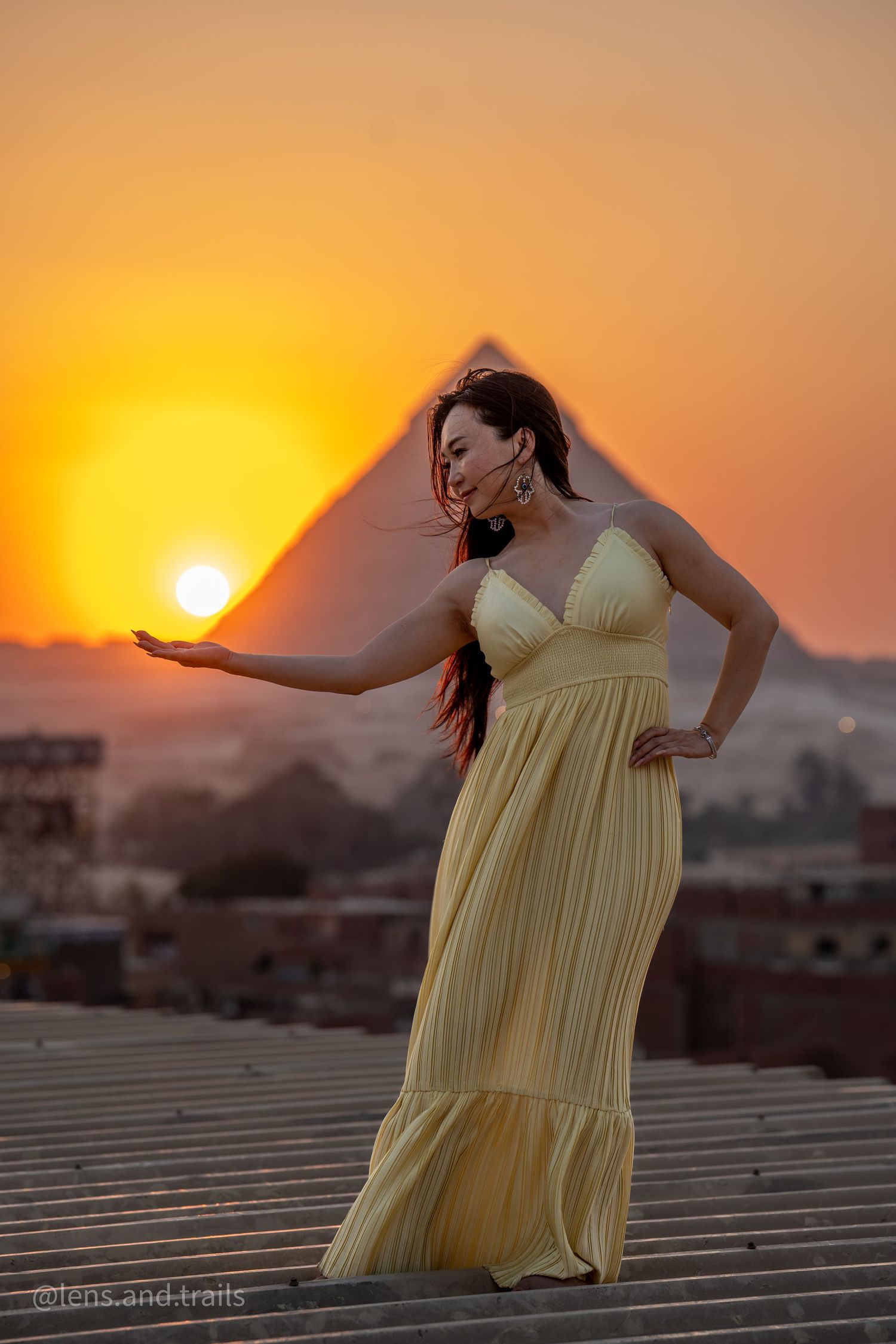 Woman in elegant yellow maxi dress poses on rooftop with Egyptian pyramid and stunning sunset behind her.