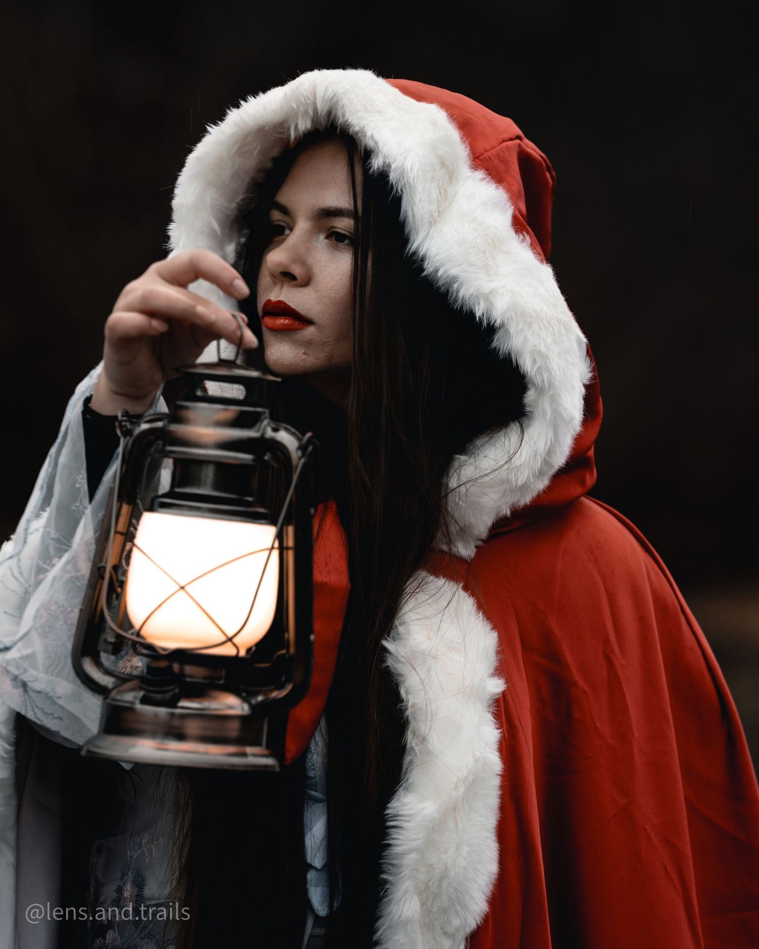 Figure in red cape with fur trim holds glowing lantern in dramatic dark setting.