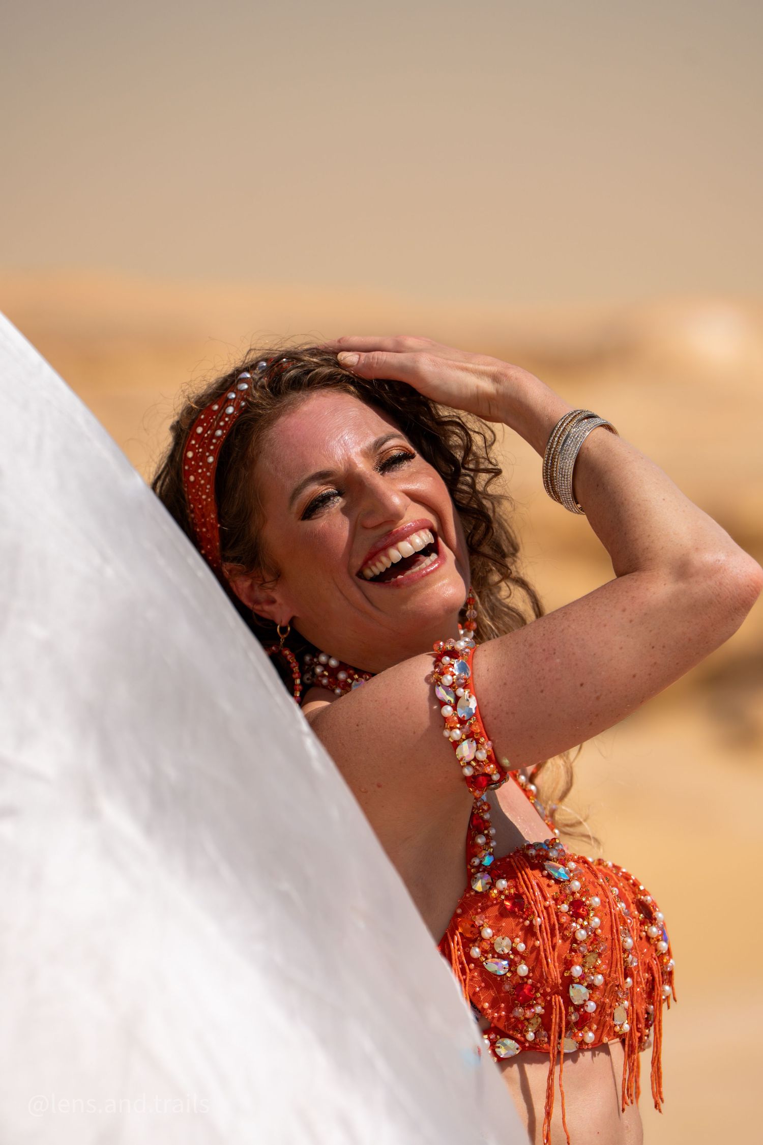 Laughing belly dancer in red beaded costume holds white veil in sandy desert environment.