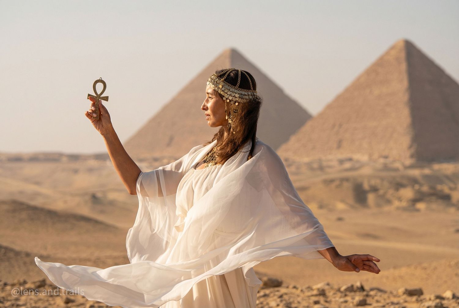 Woman in white Egyptian goddess costume holds ankh symbol before the Great Pyramids of Giza.