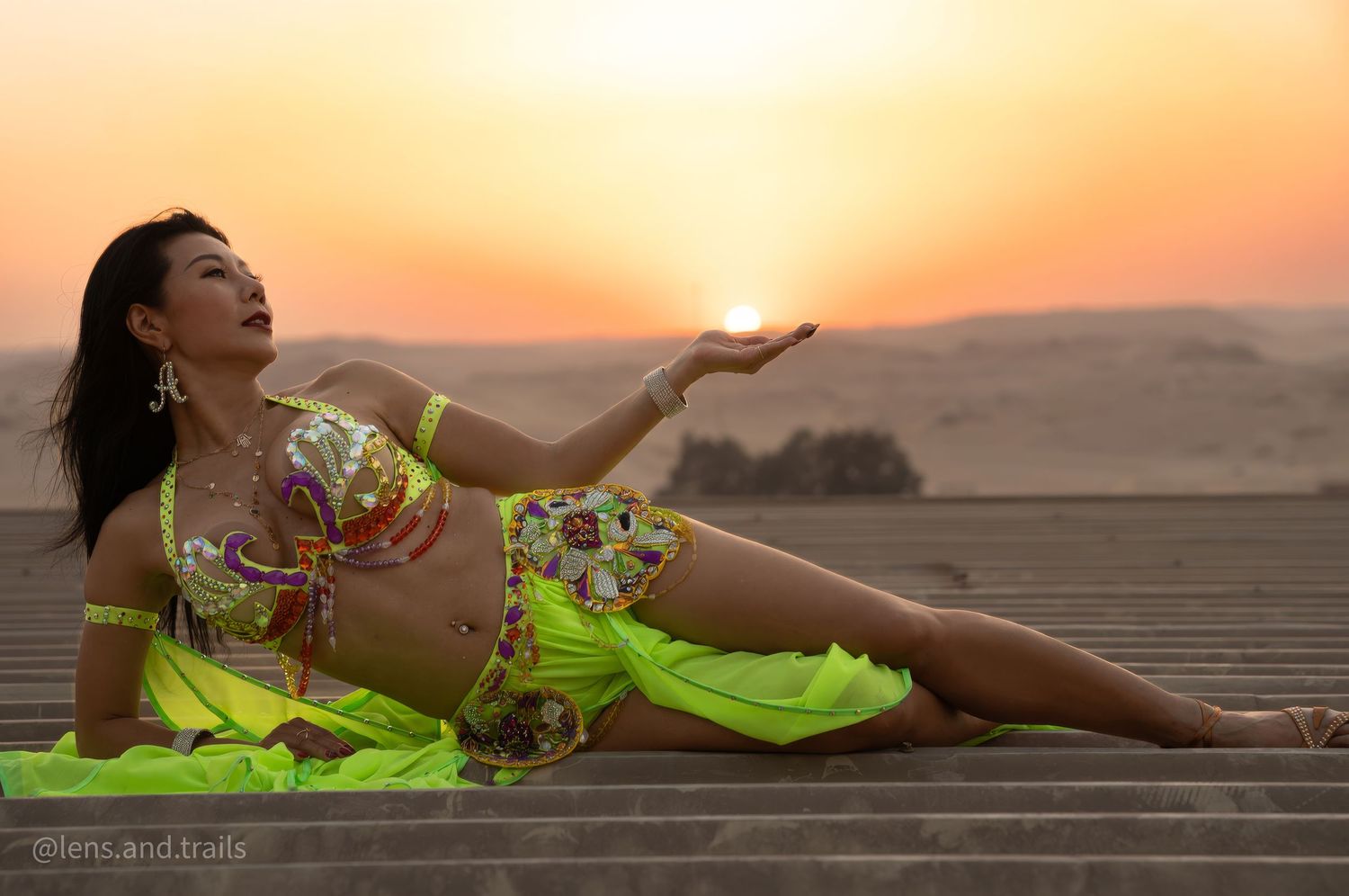 Belly dancer in neon yellow costume poses at desert sunset holding the sun.