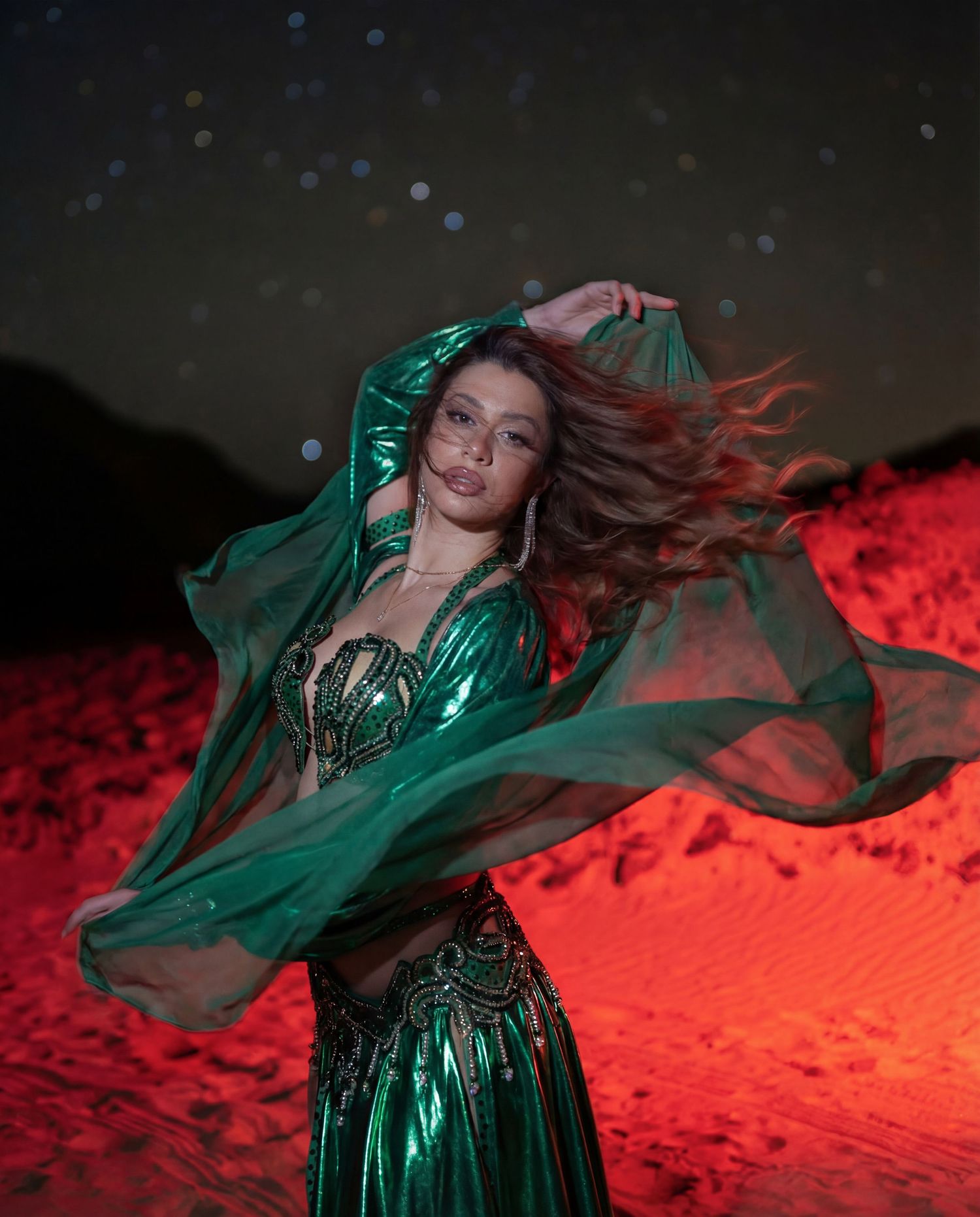 A belly dancer in emerald green costume with flowing fabric dances under a starry desert night sky.