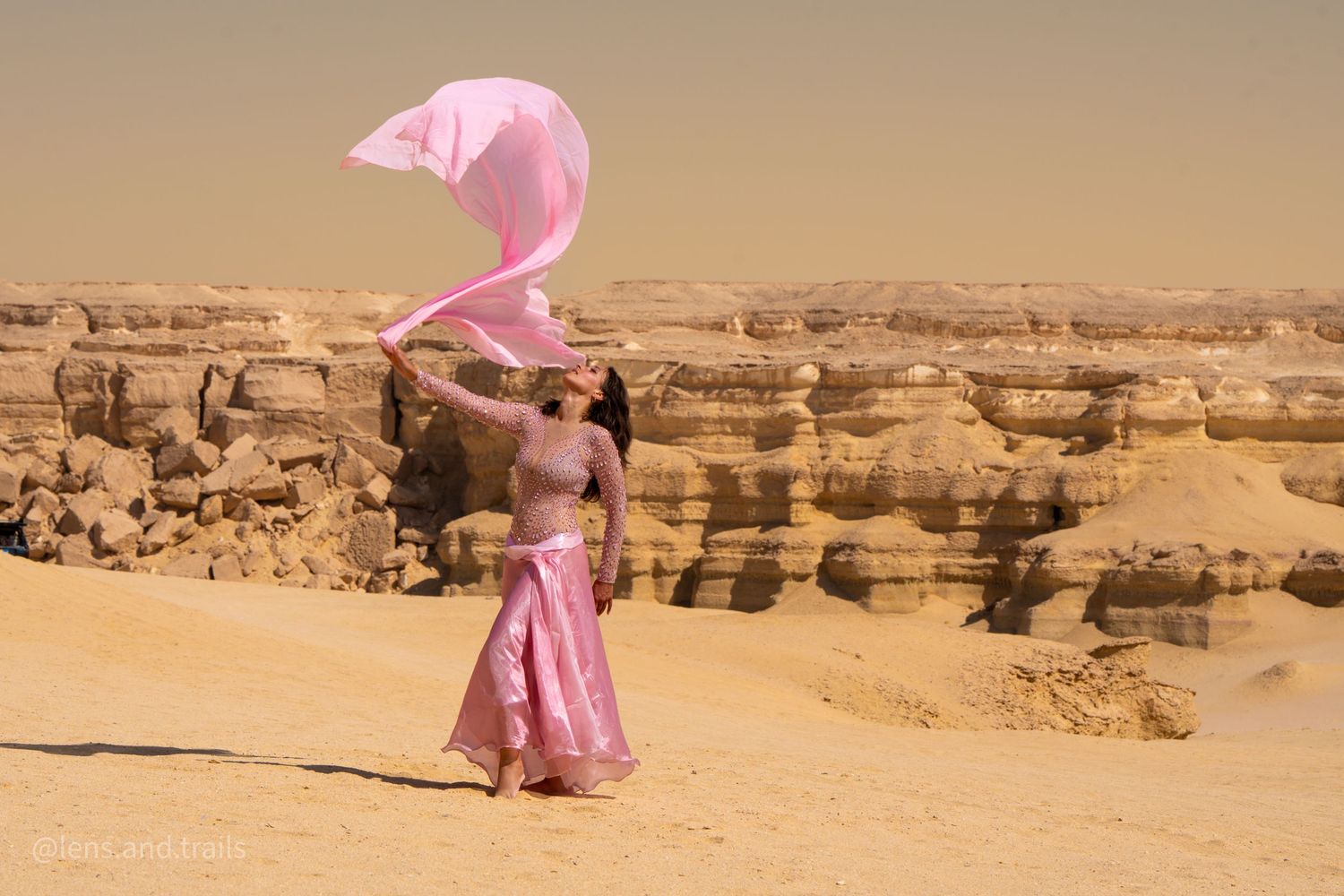 Belly dancer in pink costume twirling flowing veil in vast desert landscape with rocky cliffs.