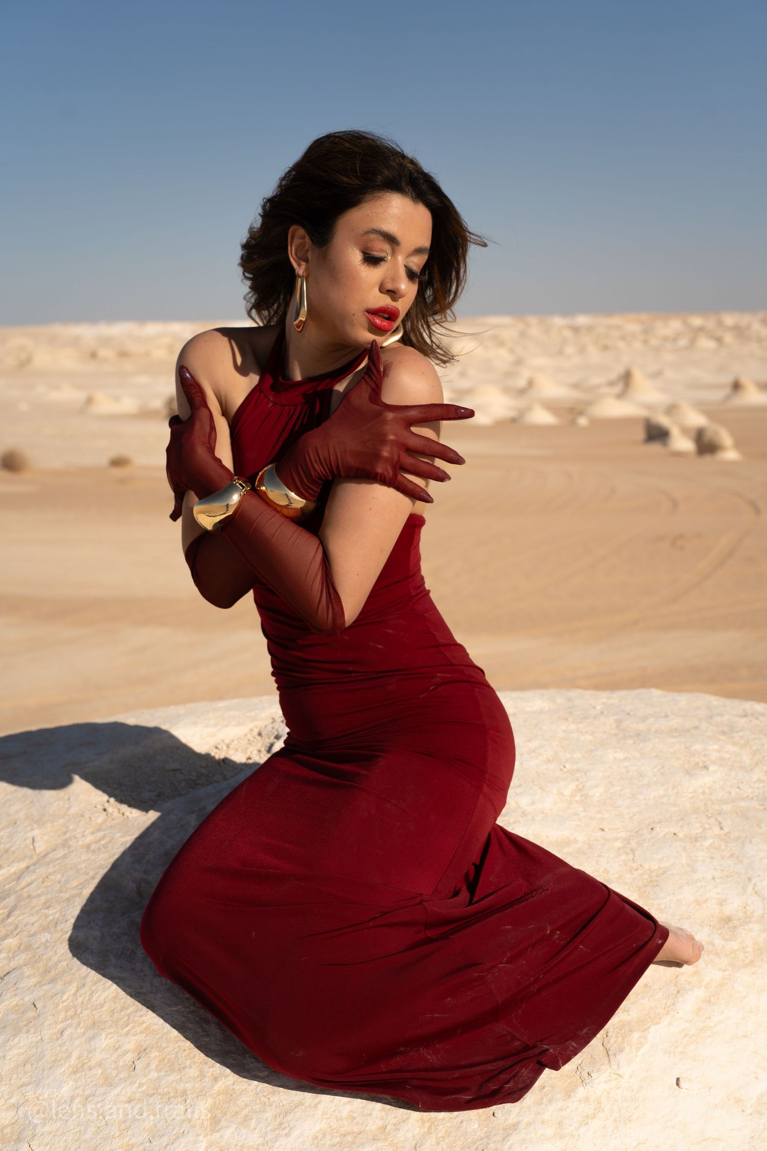 Woman in red velvet dress and gloves kneels on white desert rock surface.
