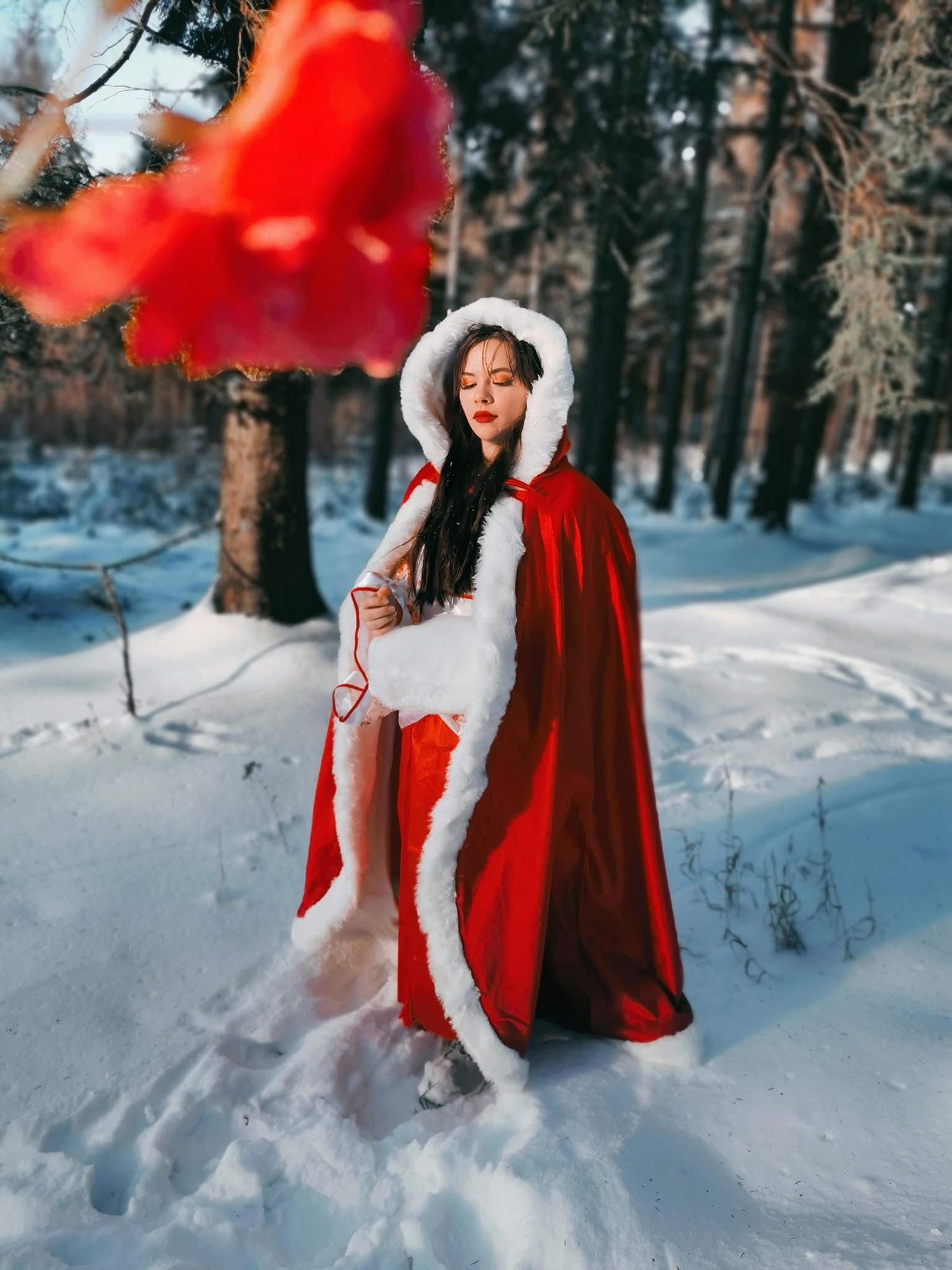 Woman in red hooded cape stands in snowy winter forest in festive holiday costume.