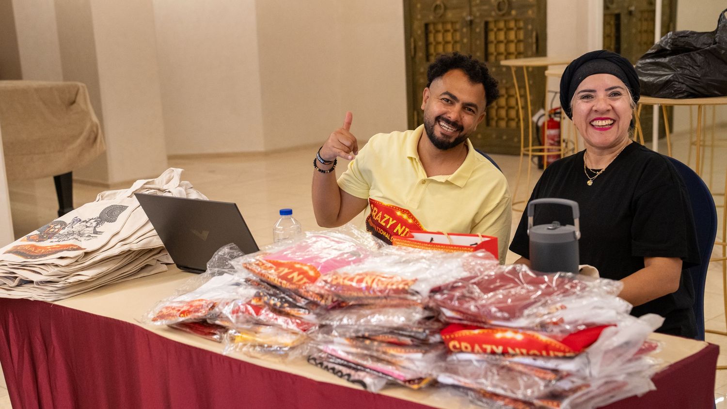 Two people smile behind a table stacked with packaged merchandise at an indoor event.