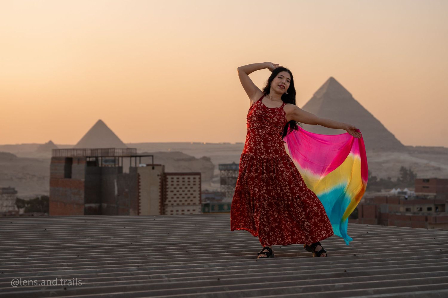 Woman in red dress dances with rainbow veil before Egyptian pyramids at dusk.