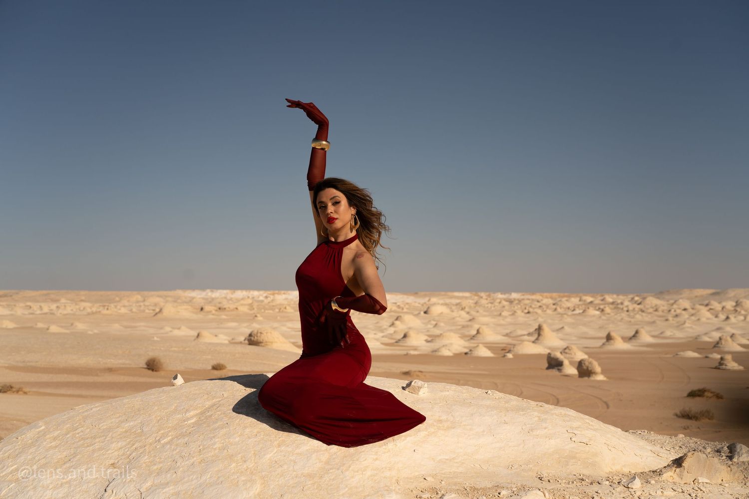Woman in red gown kneels gracefully on white sand dunes in vast desert landscape.