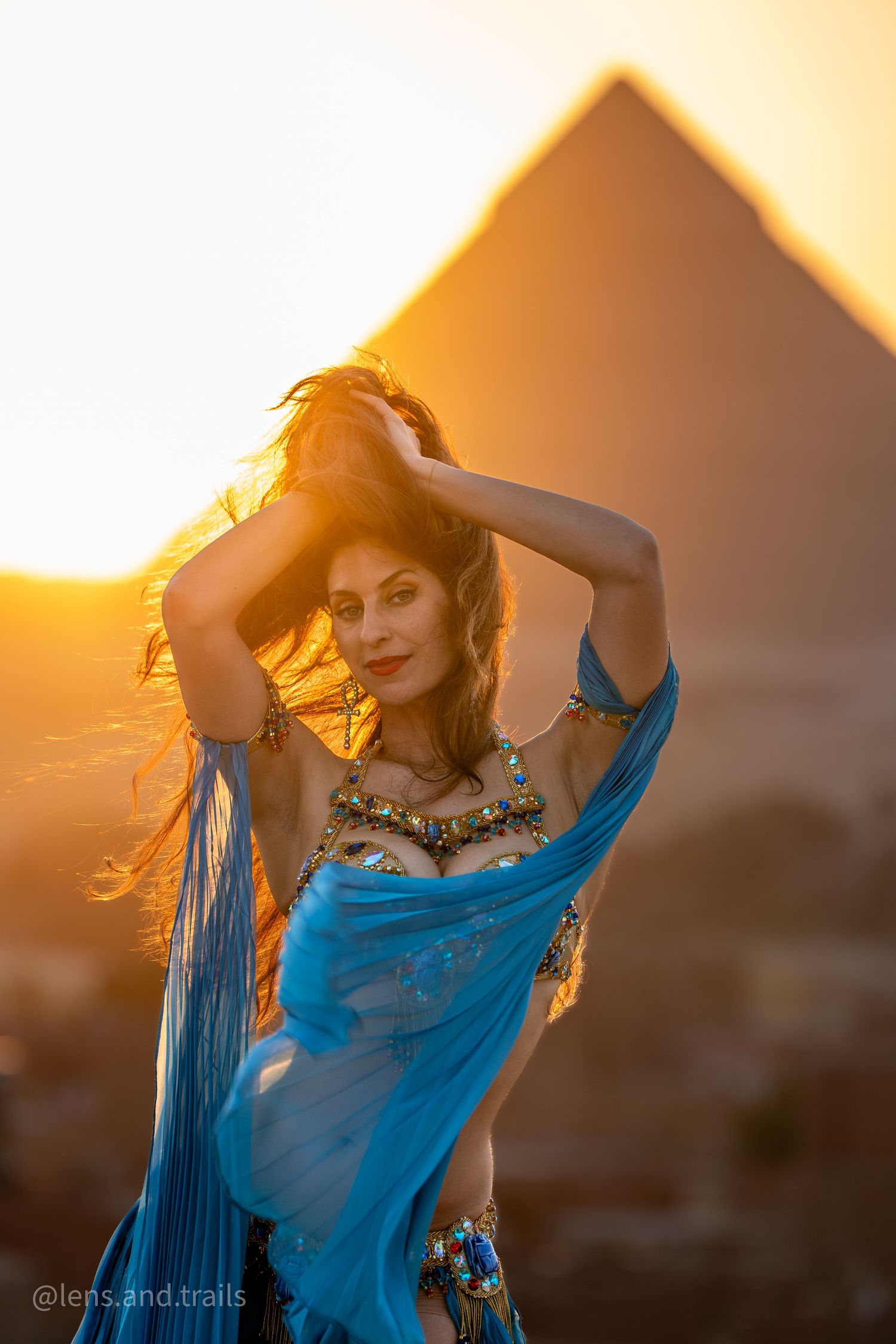 Woman in blue belly dance costume poses before Egyptian pyramid at golden hour.