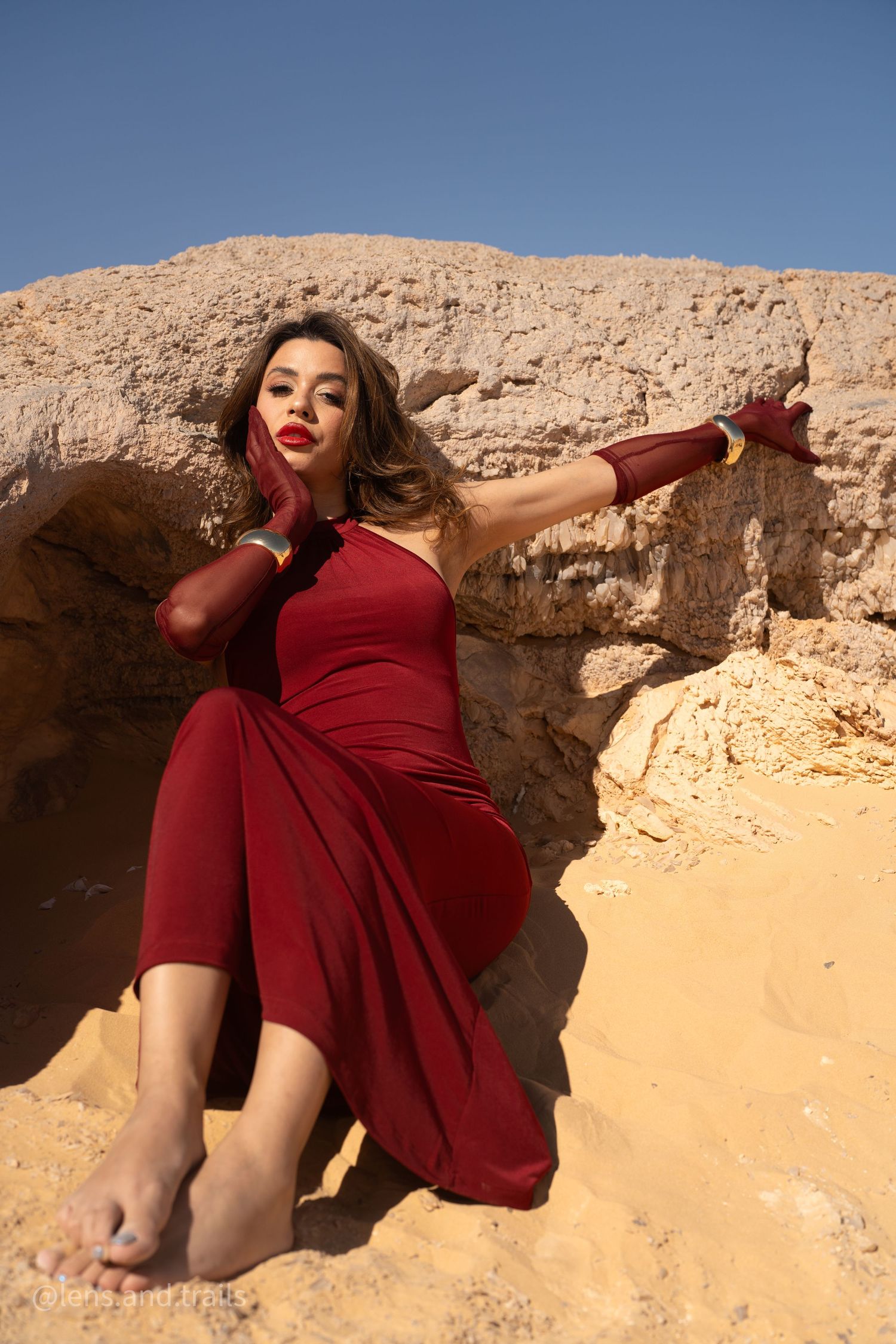Woman in red dress and gloves sits gracefully against sandy desert rock wall.
