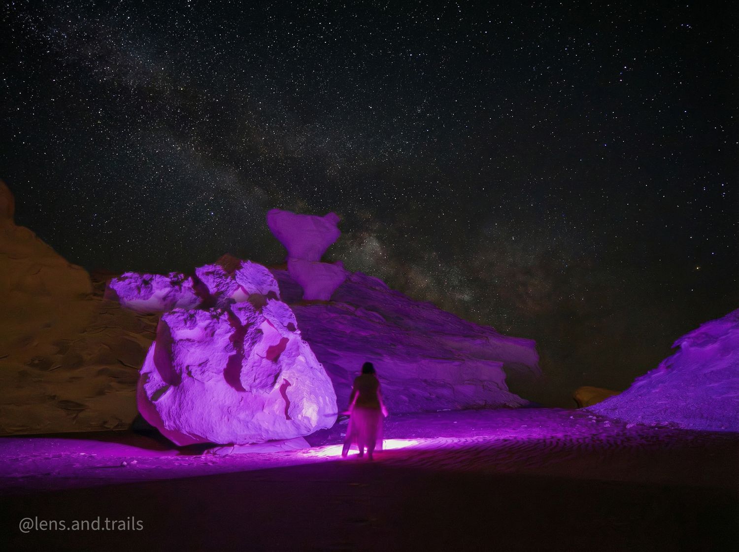 Lone figure glows in purple light beside massive desert rock formations under the Milky Way.