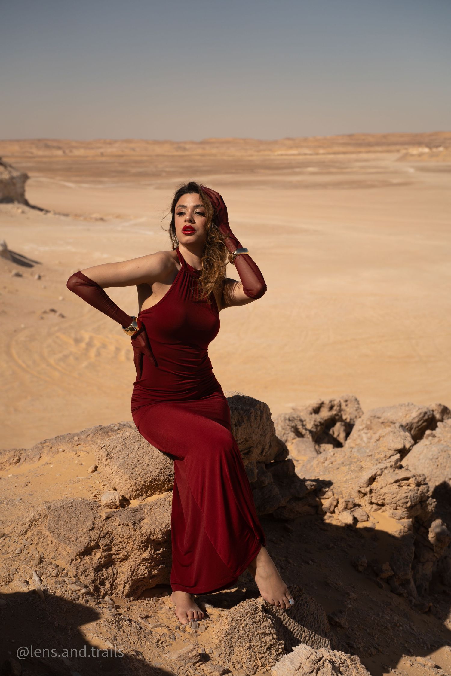 Woman in red gown poses on desert rocks with vast sandy landscape behind her.