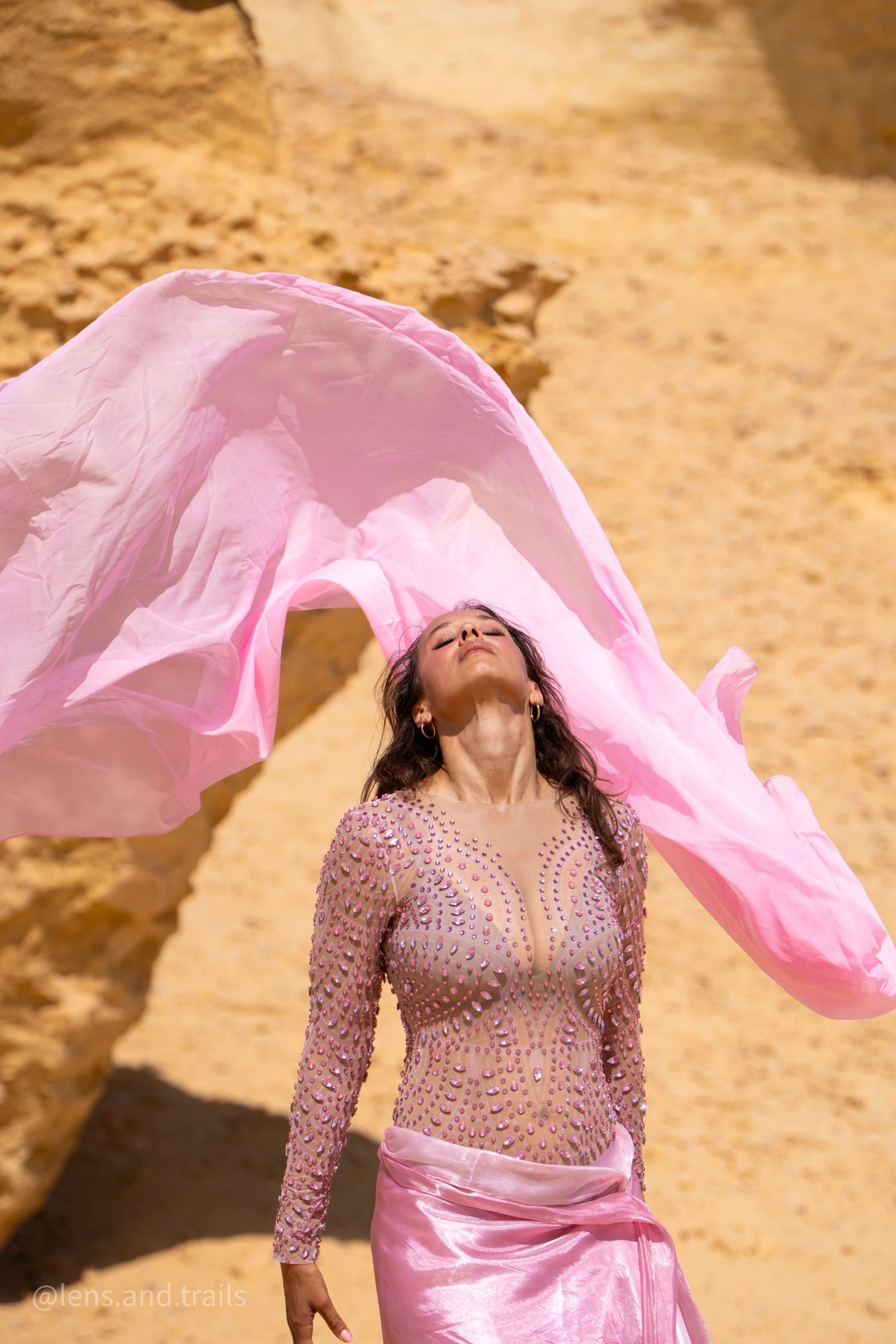 Belly dancer in beaded costume tosses flowing pink silk veil upward in sunlit desert canyon.