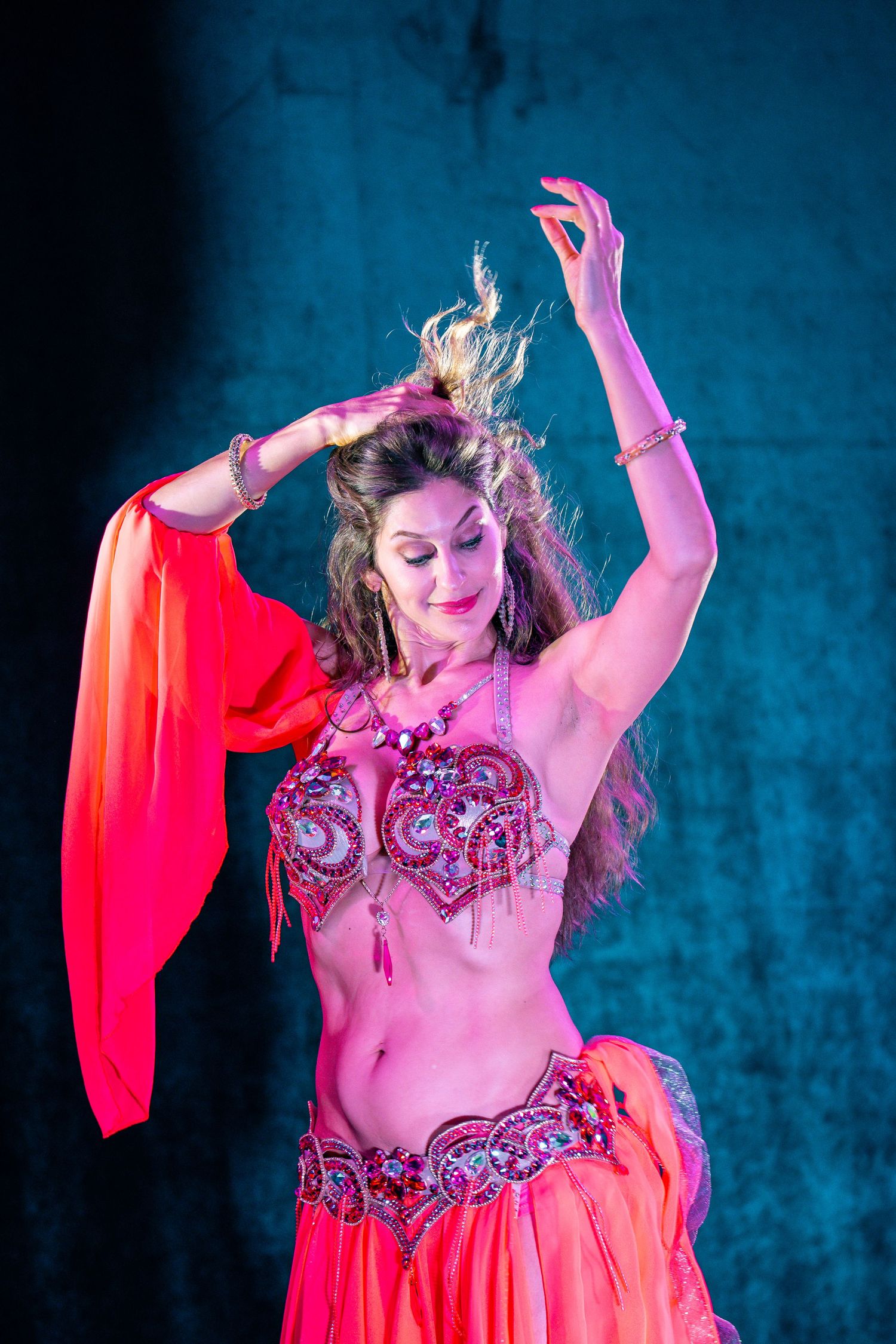 A belly dancer in a pink embellished costume performs with flowing red veils under dramatic lighting.