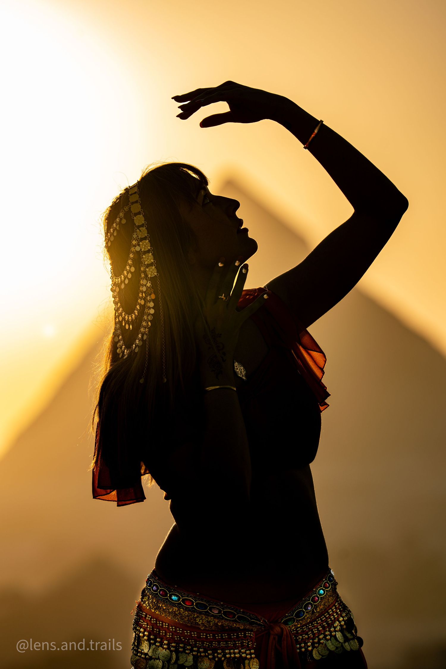 Silhouette of a belly dancer with traditional headpiece and raised hand against pyramid sunset backdrop.