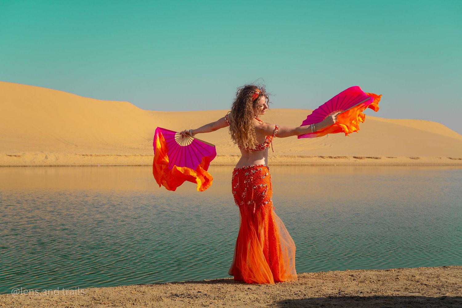 Belly dancer in orange costume holds colorful fans near desert lake with sand dunes and clear sky.
