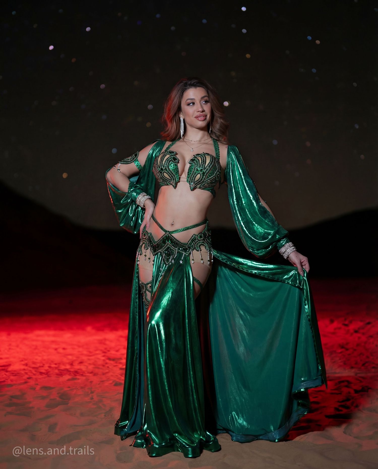 Woman in emerald green belly dance costume stands on red-lit desert sand at night.