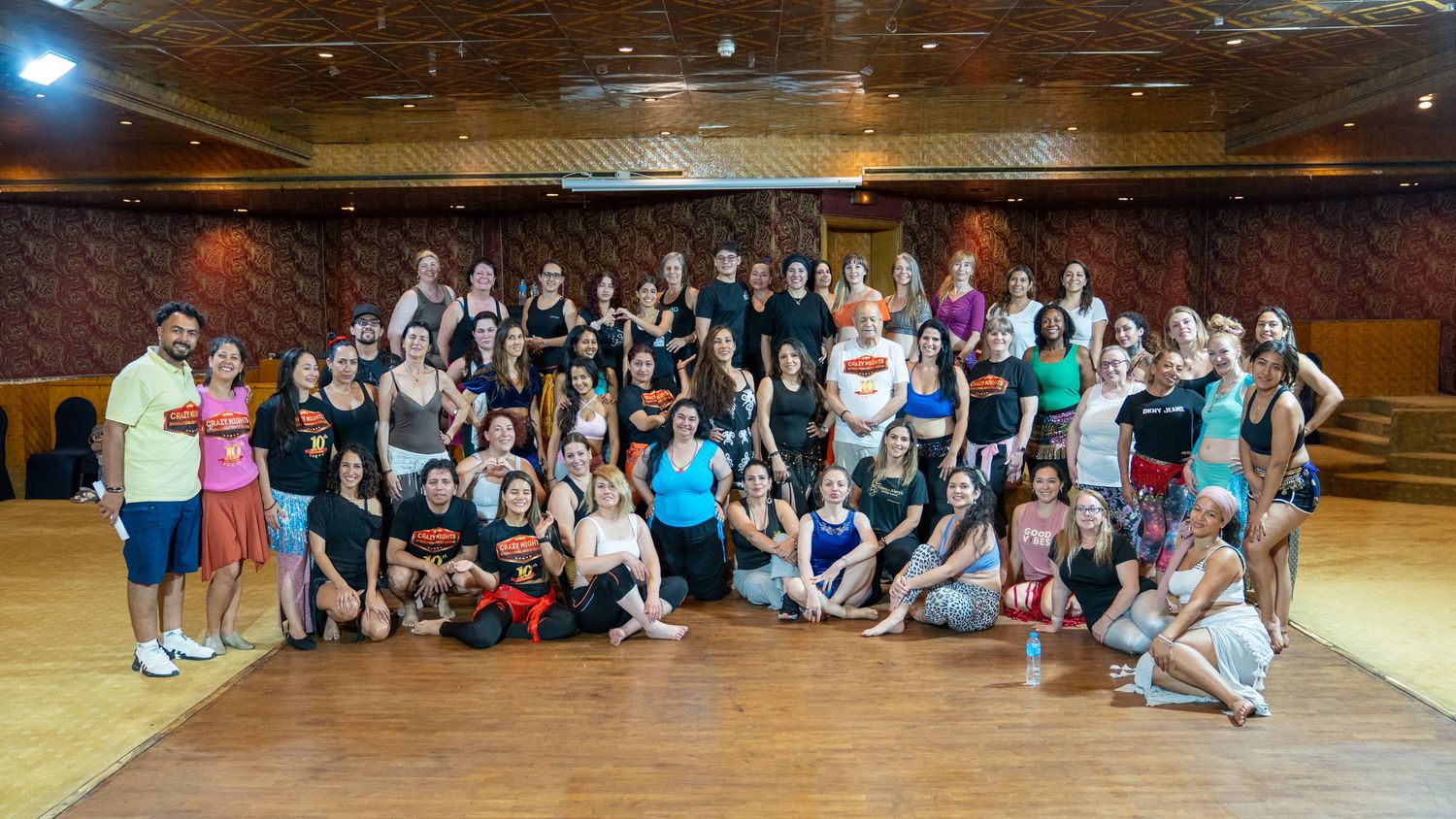 Large group of dancers poses together in a spacious mirrored dance studio.