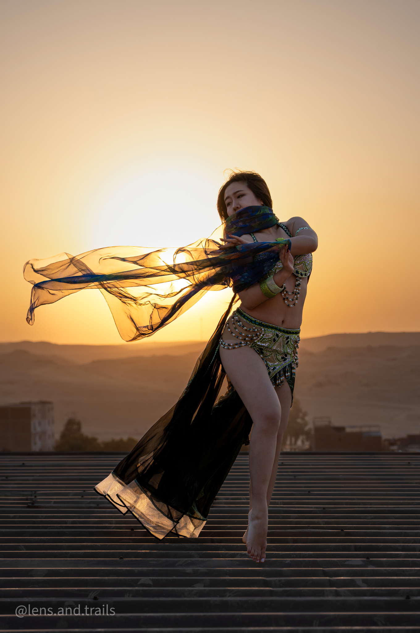 Belly dancer in black costume twirling sheer veil on rooftop at golden sunset.