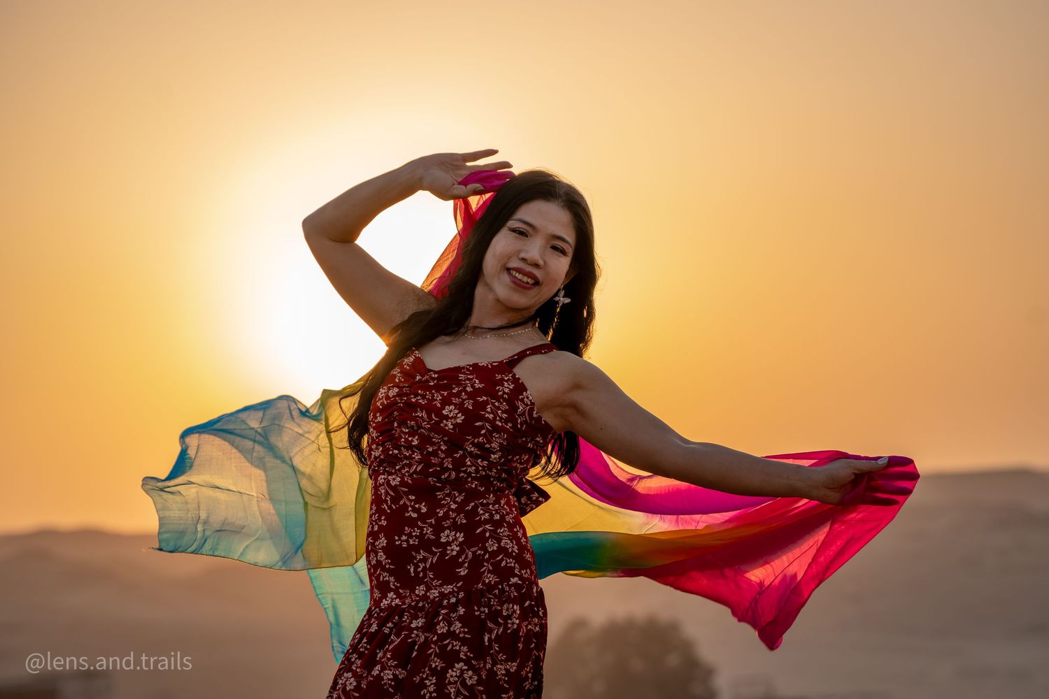 Smiling woman in red dress posing with rainbow silk scarves against golden sunset sky.
