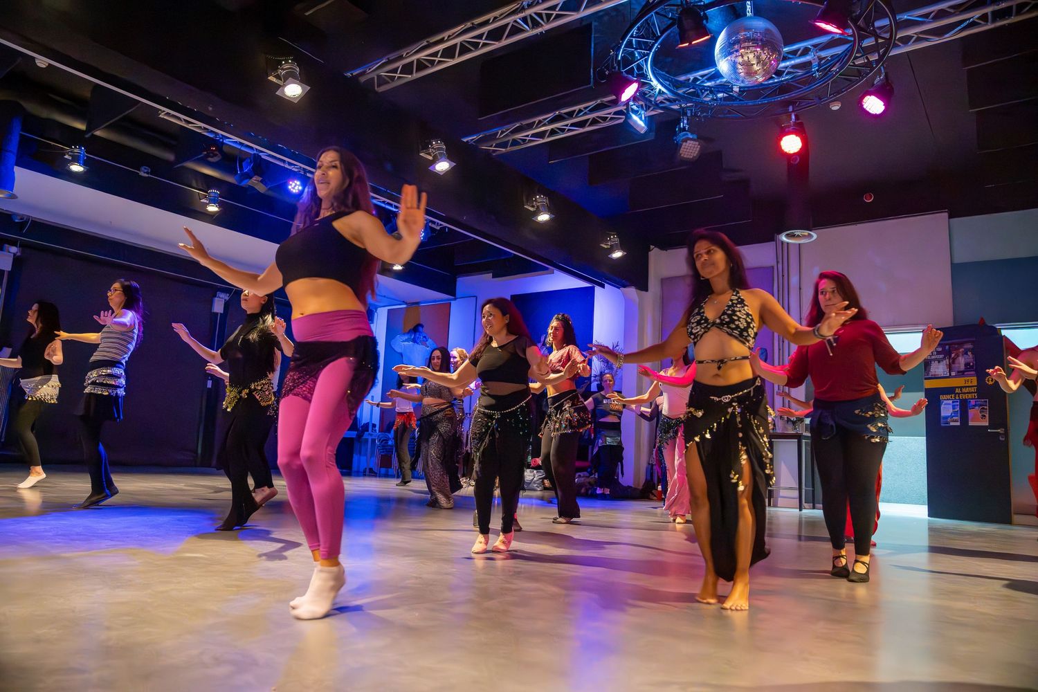 Energetic belly dance class with women in colorful costumes practicing moves in a neon-lit dance studio.