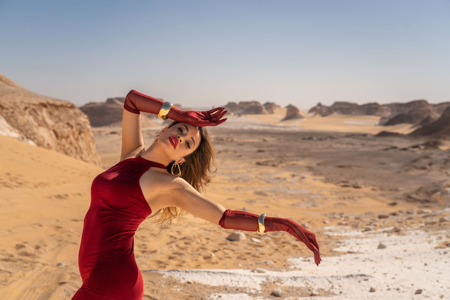 Woman in red dress and burgundy gloves dances gracefully in sunny Egyptian desert landscape.