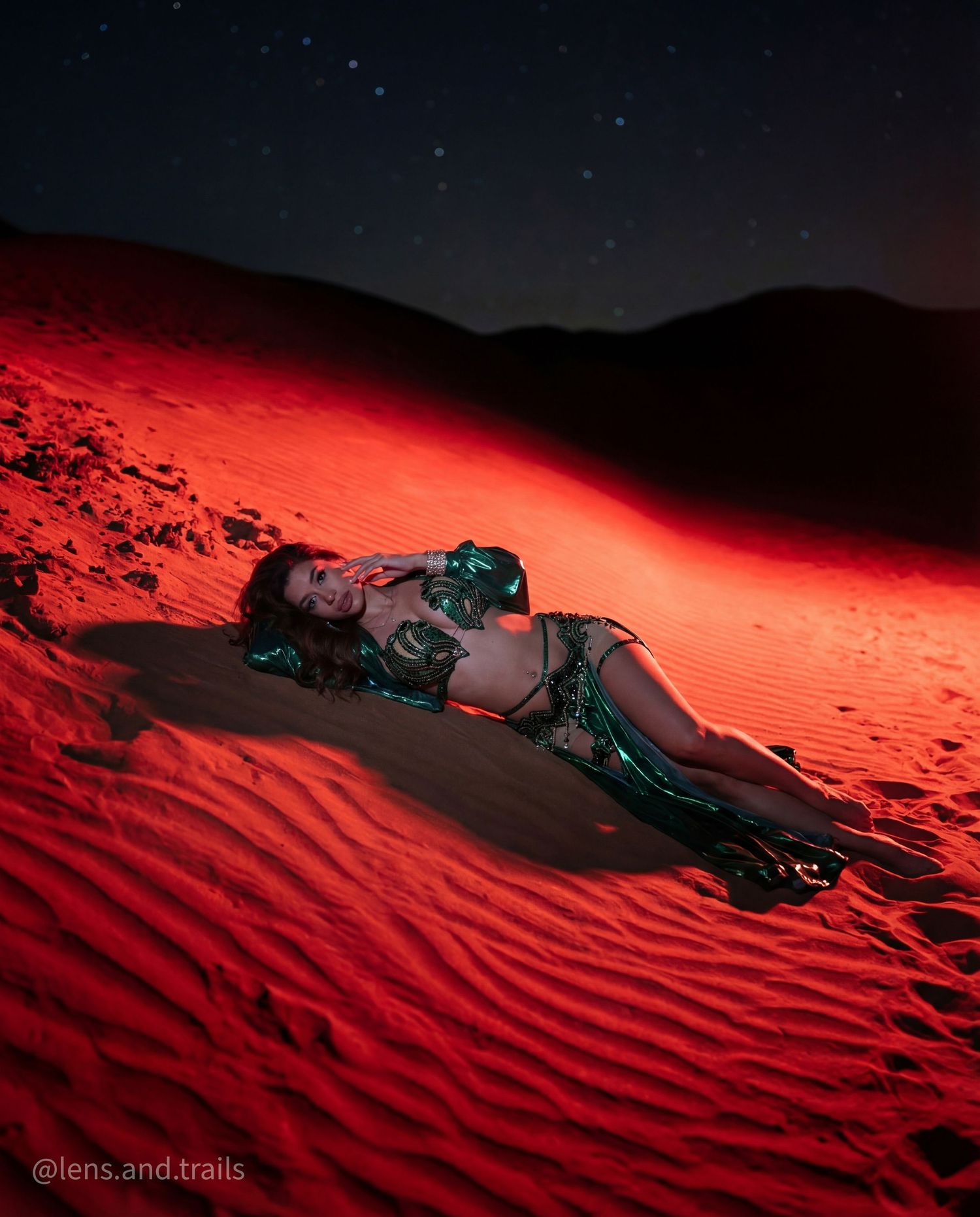 Belly dancer in green costume reclines on red-lit rippled desert sand dunes under stars.