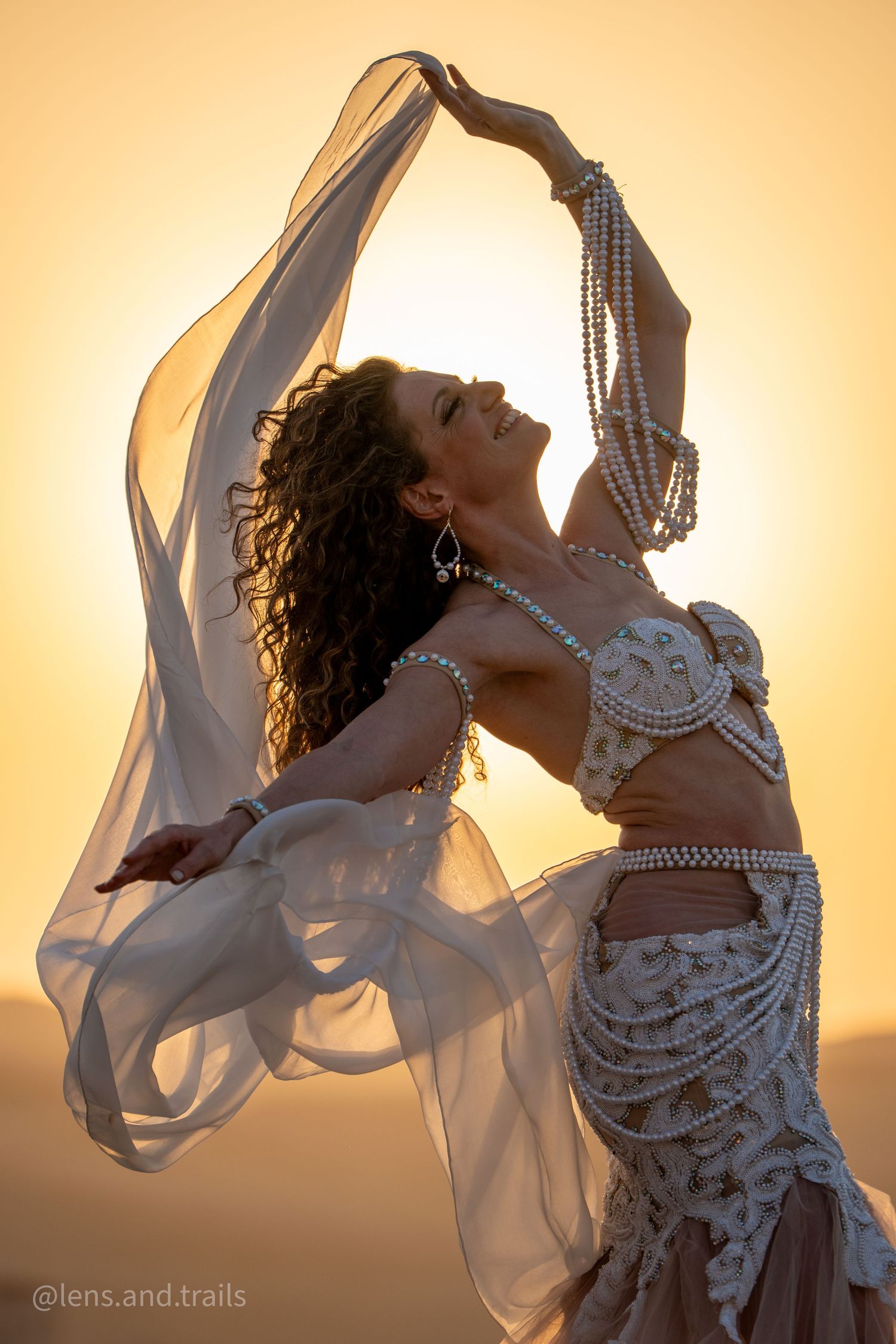 A belly dancer in white pearl costume holds a veil overhead against a golden desert sunset sky.