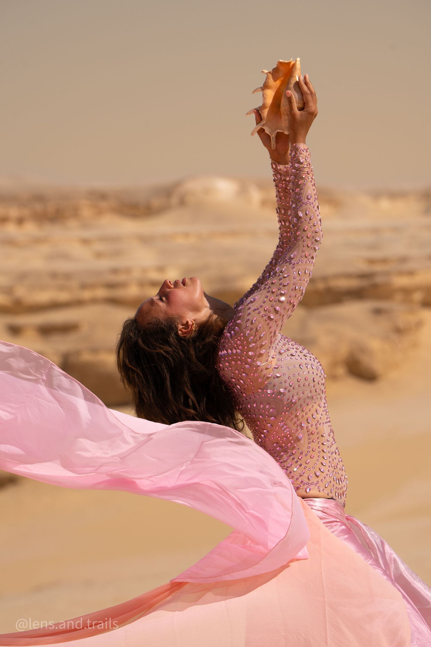 Woman in pink embellished dress reaching seashell skyward amid flowing pink fabric in desert.