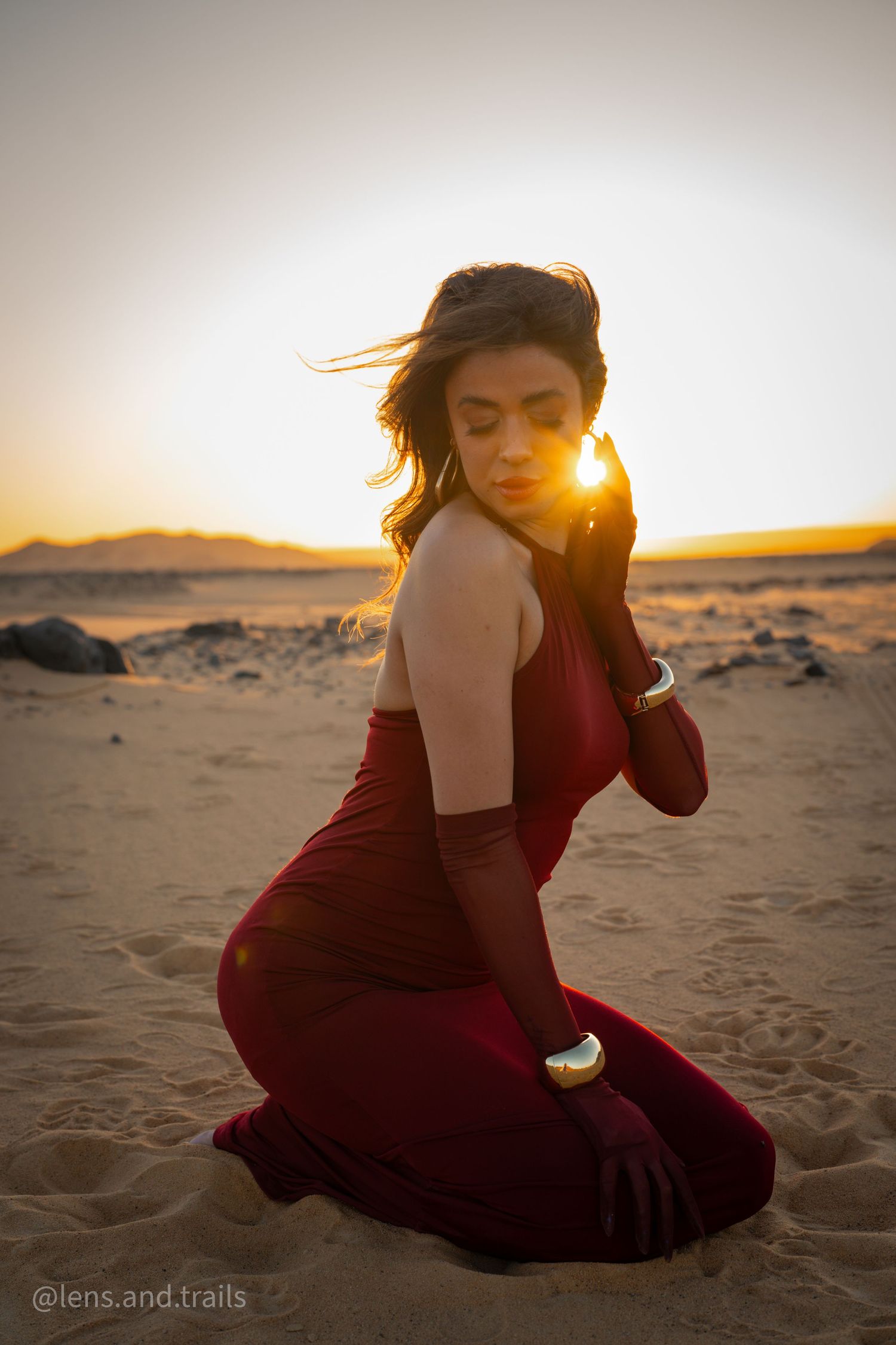 Silhouetted woman in red dress kneels on sand dunes during golden desert sunset.