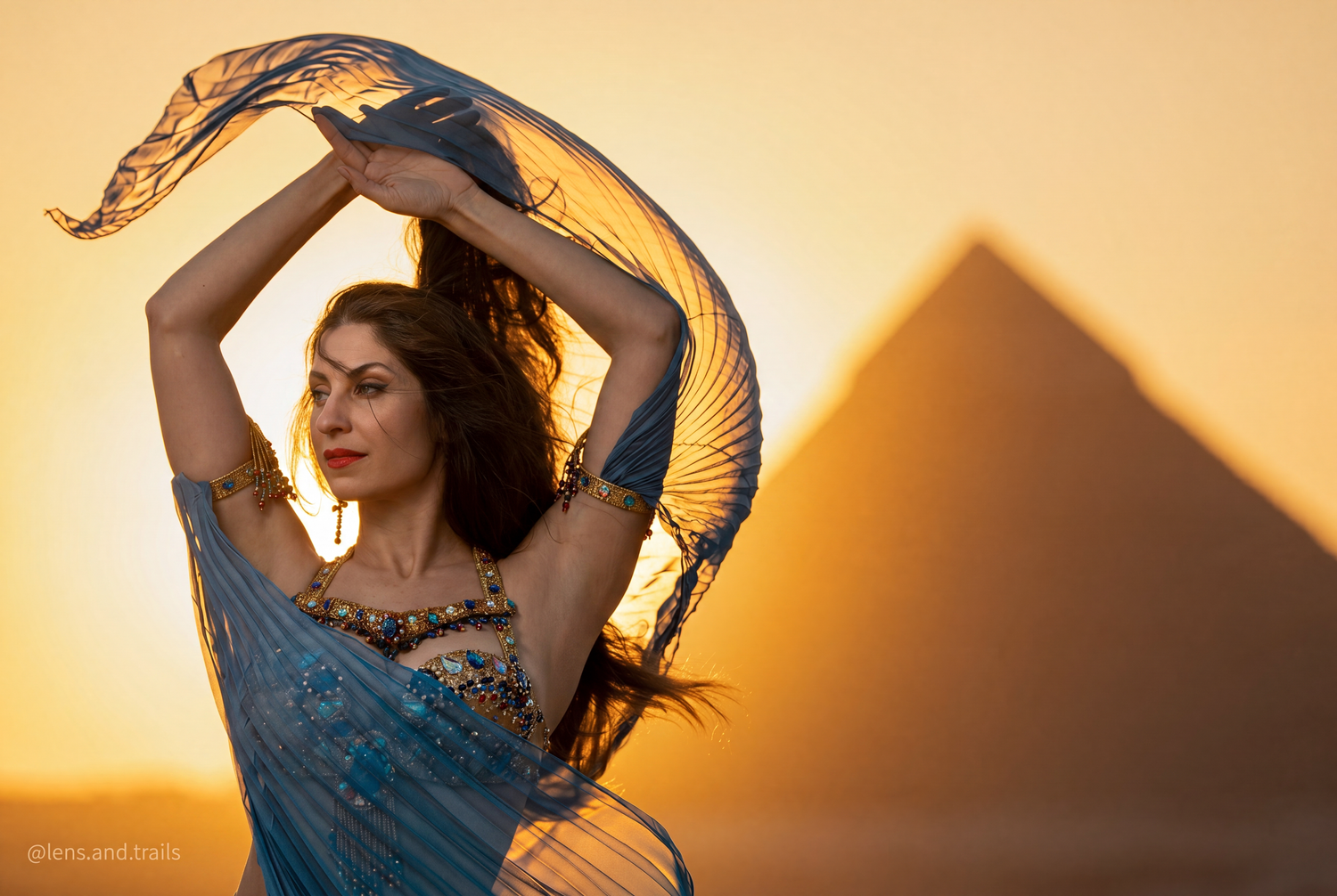 A belly dancer in a blue costume performs gracefully at sunset with the Egyptian pyramids in the background.