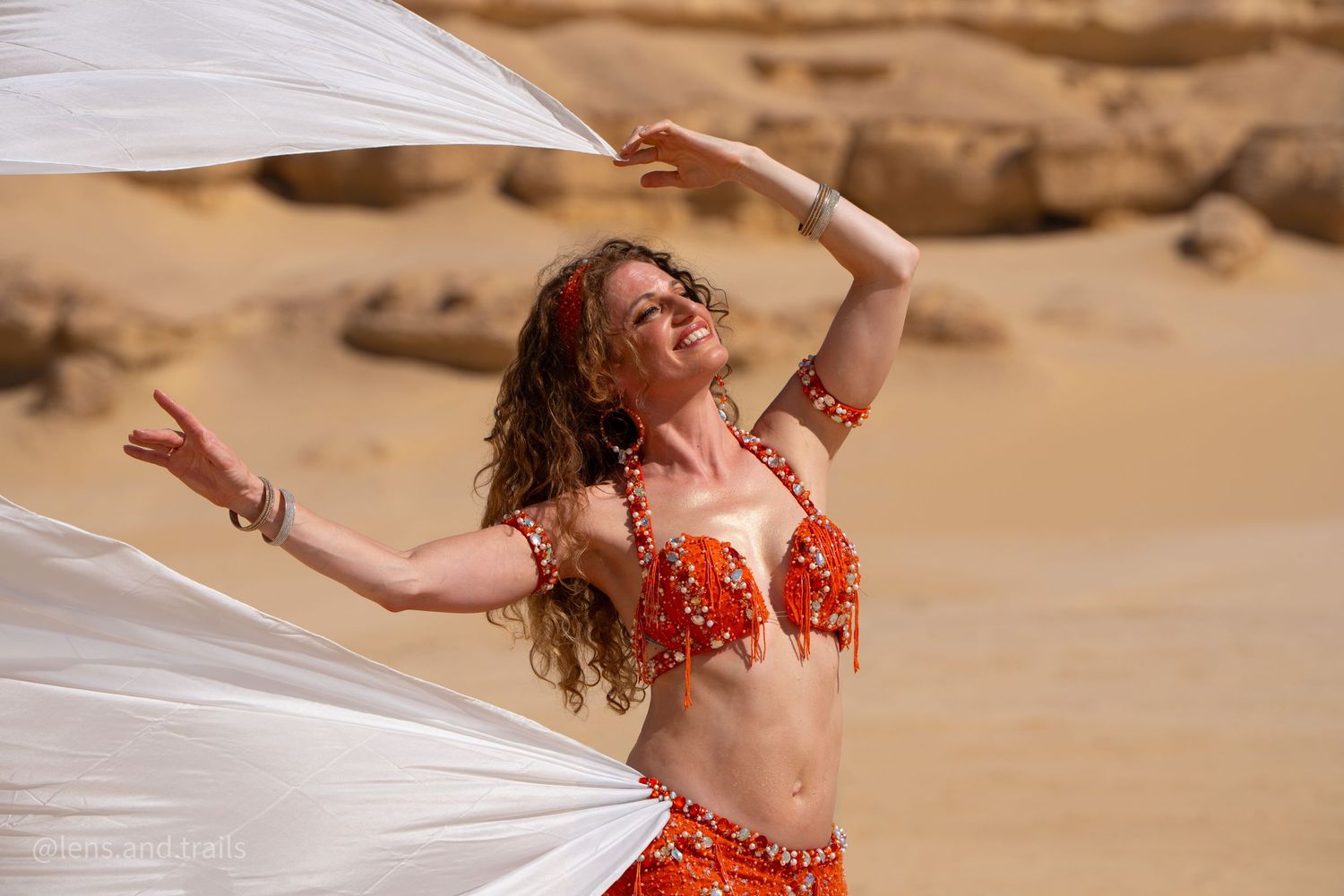 Belly dancer in red beaded bra and skirt poses with white flowing fabric in desert rocky setting.