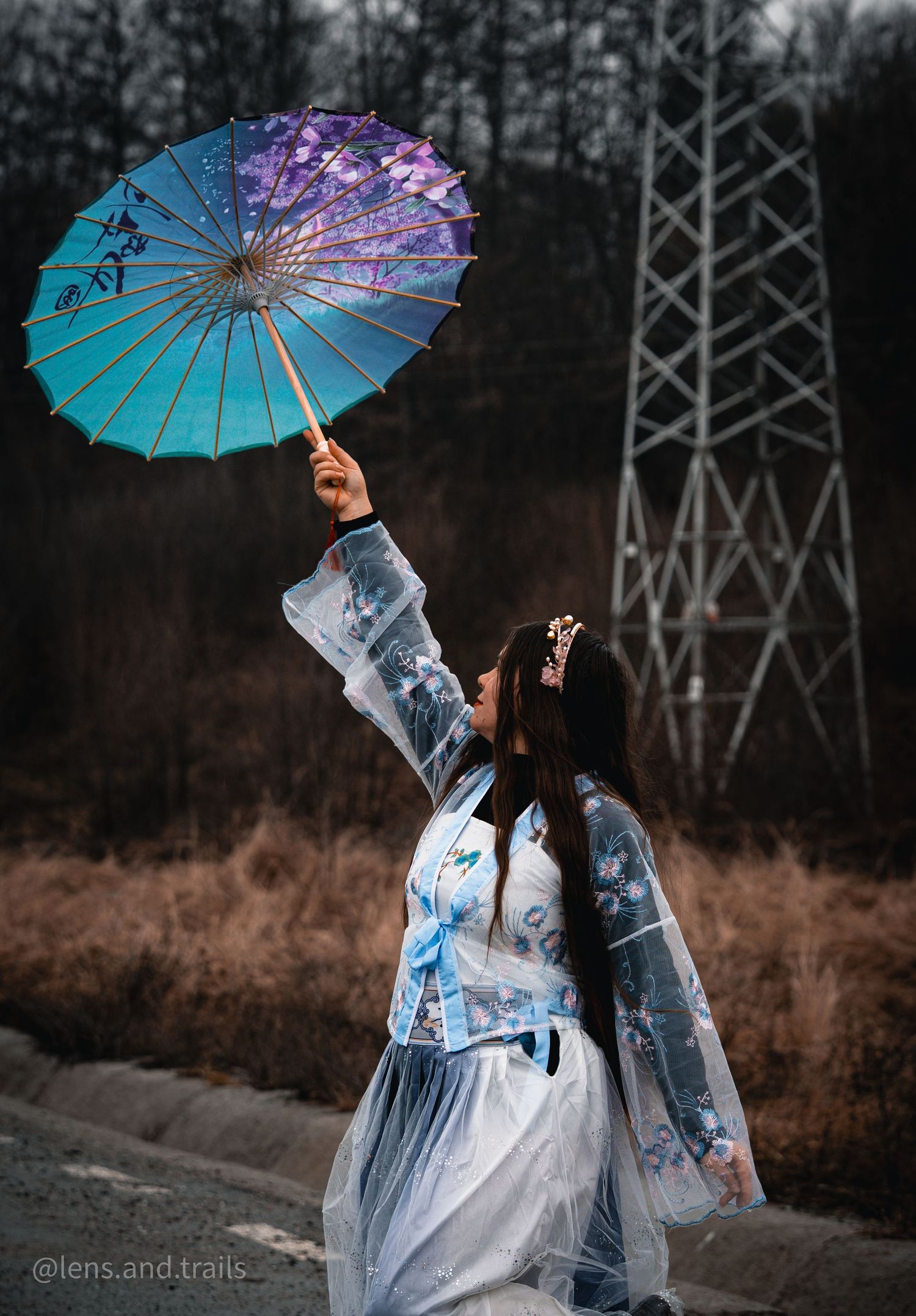 Figure in flowing white dress twirls blue parasol near power lines at dusk.