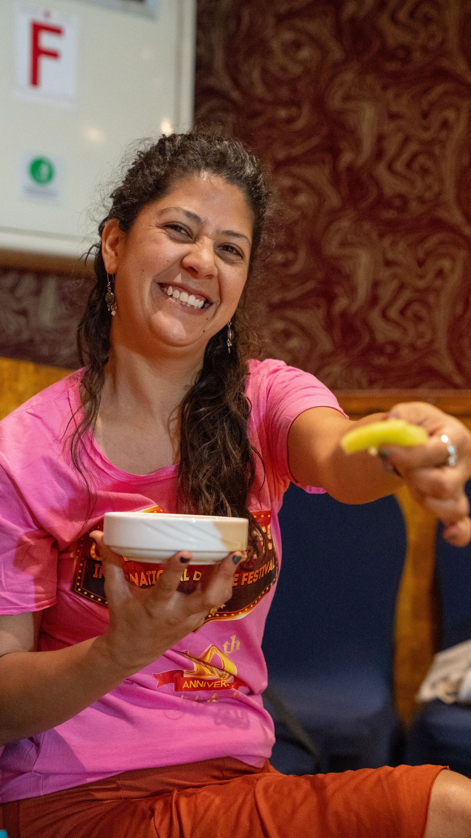 Smiling woman in pink shirt holds a bowl and extends a banana toward the camera indoors.