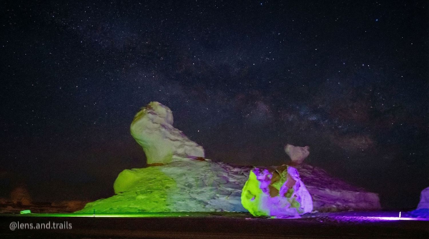 Desert rock formations glow in vibrant purple and green lights under a brilliant Milky Way sky.