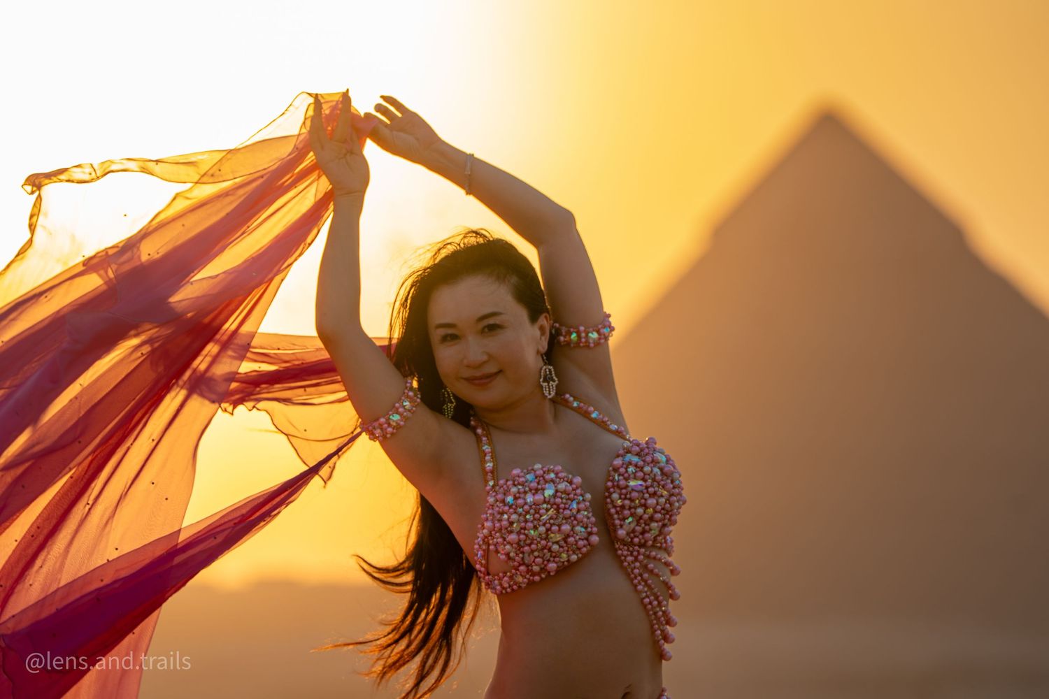 Belly dancer in jeweled costume with red veil poses before Egyptian pyramids at golden sunset.