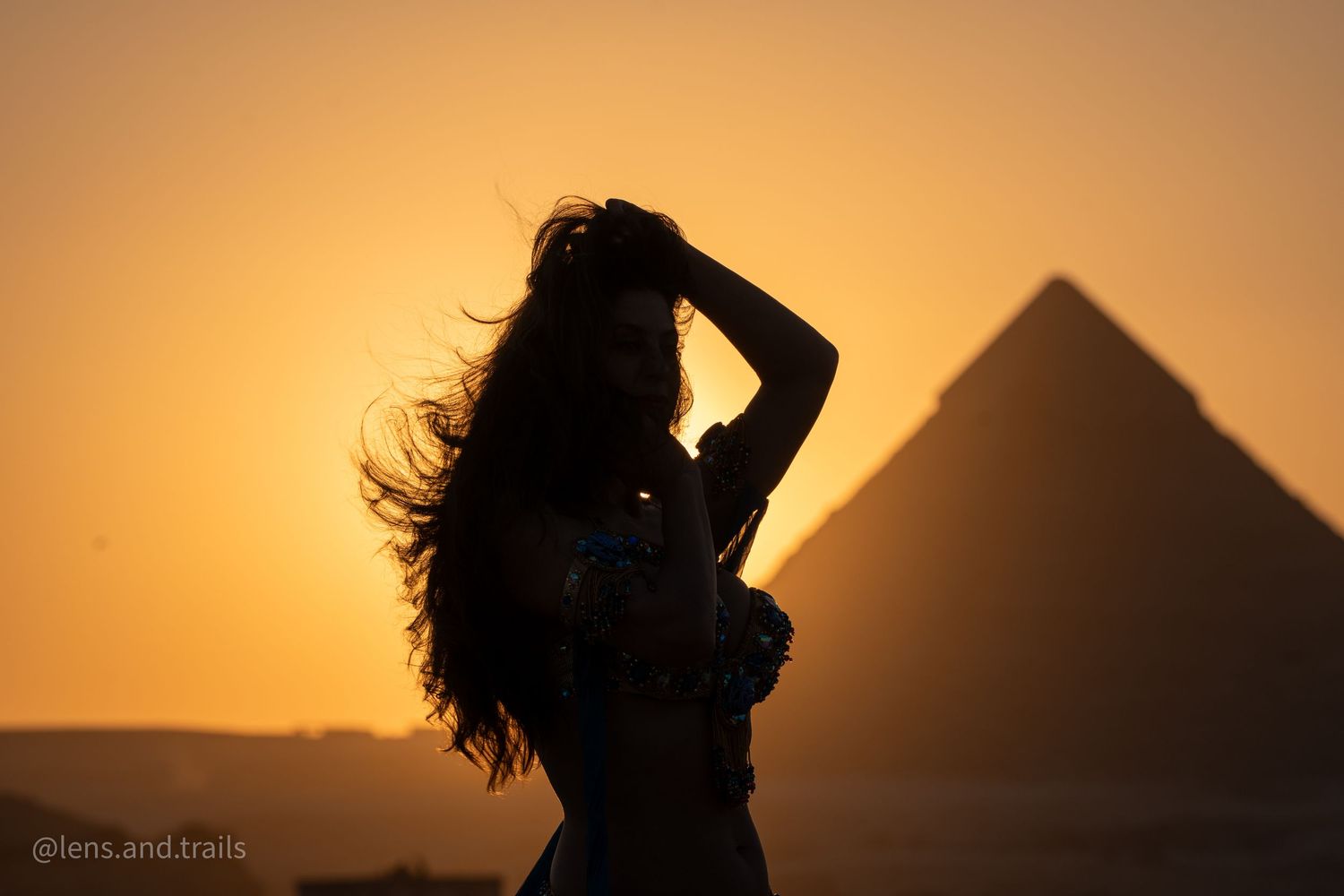 Silhouette of woman with long hair posing before Egyptian pyramid at sunset.
