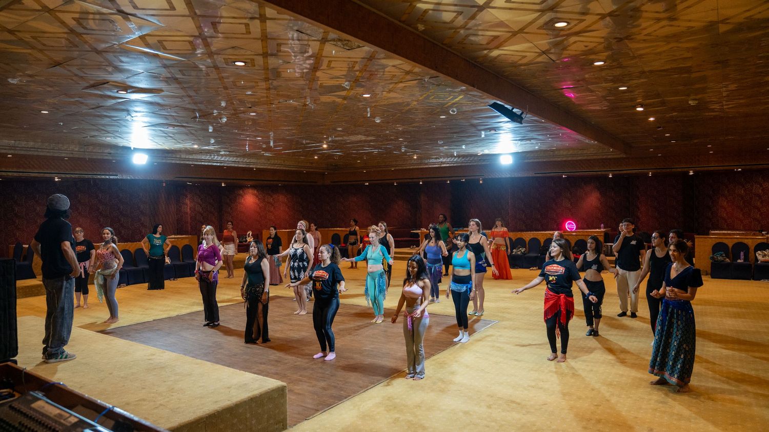 Large group of dancers practice belly dance choreography in spacious wooden-floored dance studio.
