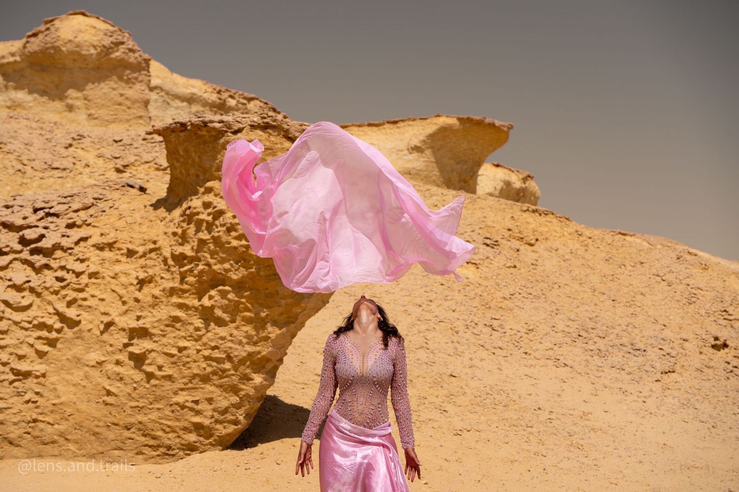 Dancer in pink belly dance costume tosses flowing pink veil in golden desert canyon setting.