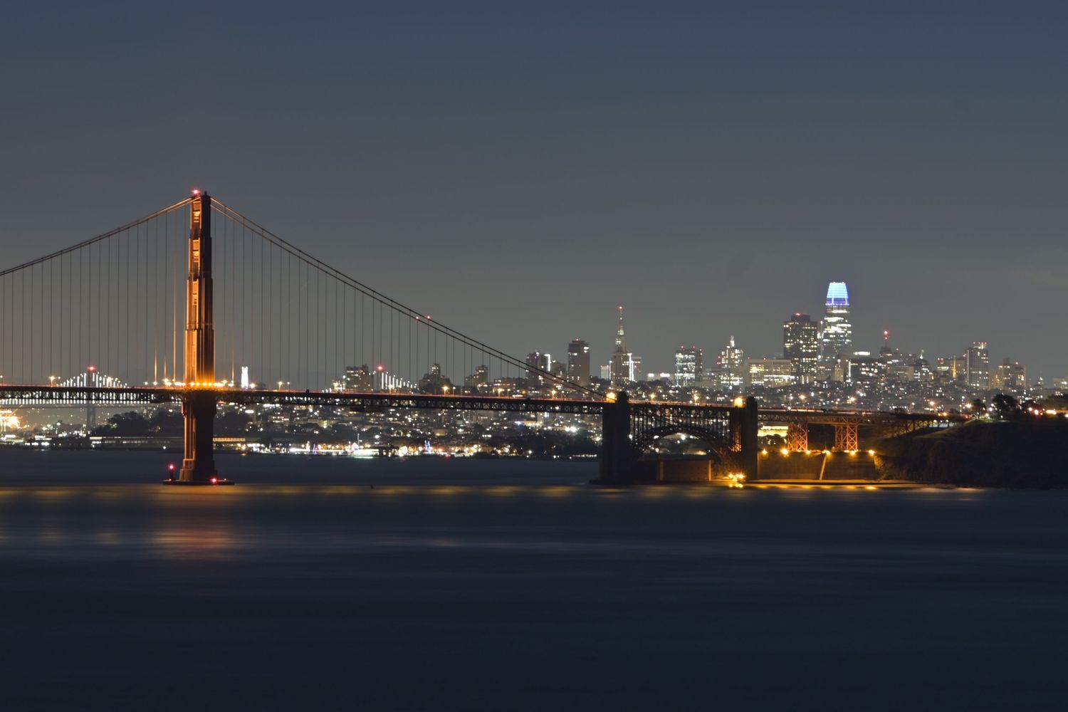 Golden Gate Bridge illuminates San Francisco skyline at night with city lights reflecting on the bay.